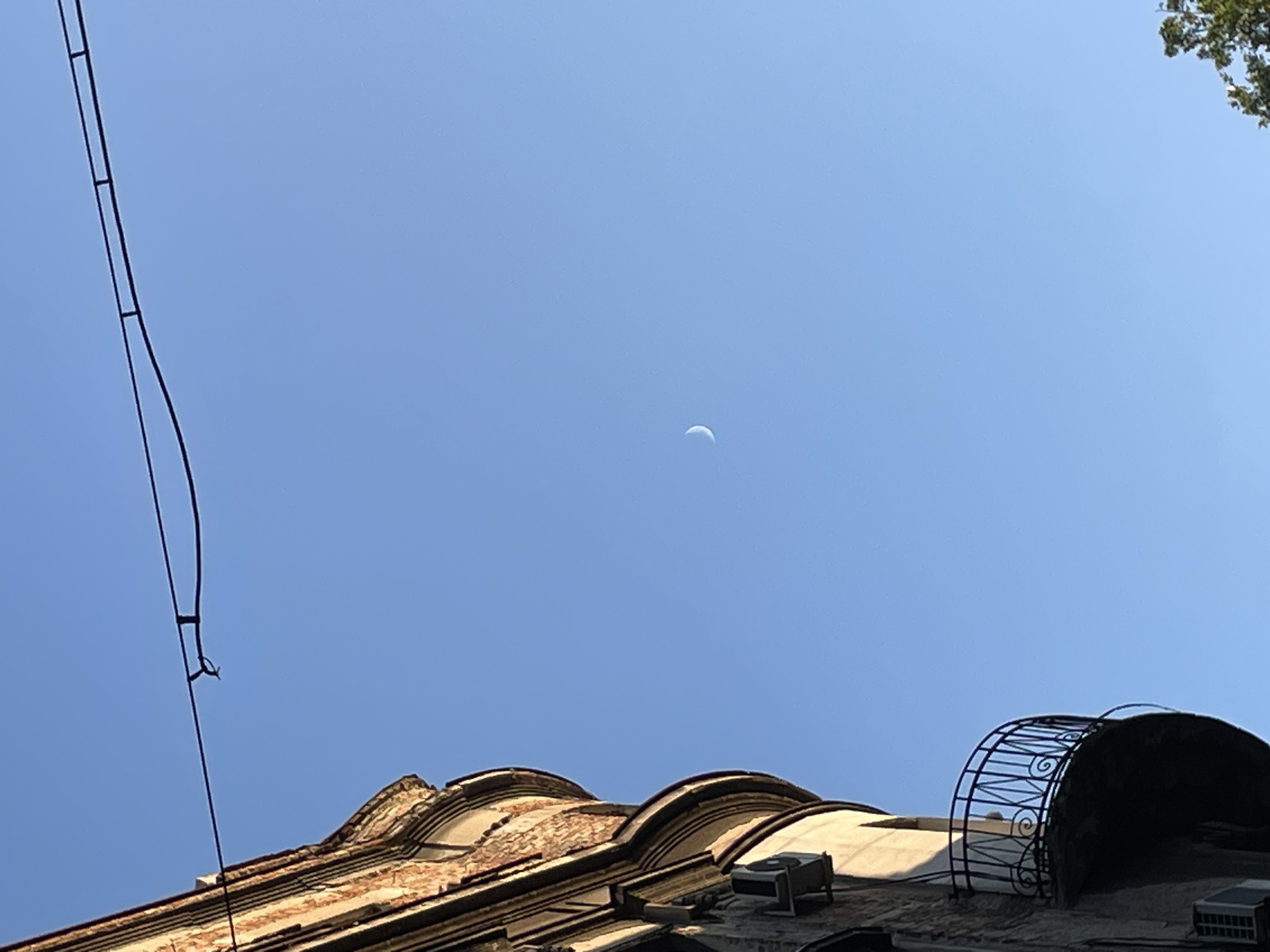 The image captures a view of a crescent moon in a clear blue sky, partially obscured by overhead wires. To the left, a section of an old, ornate building is visible, featuring weathered brickwork and a curved balcony with a black metal railing. Air conditioning units are mounted on the building's facade. A sliver of greenery from a tree is visible in the lower right corner. This scene is characteristic of the Dorćol neighborhood in Belgrade, Serbia, known for its historic architecture and charming residential streets.
