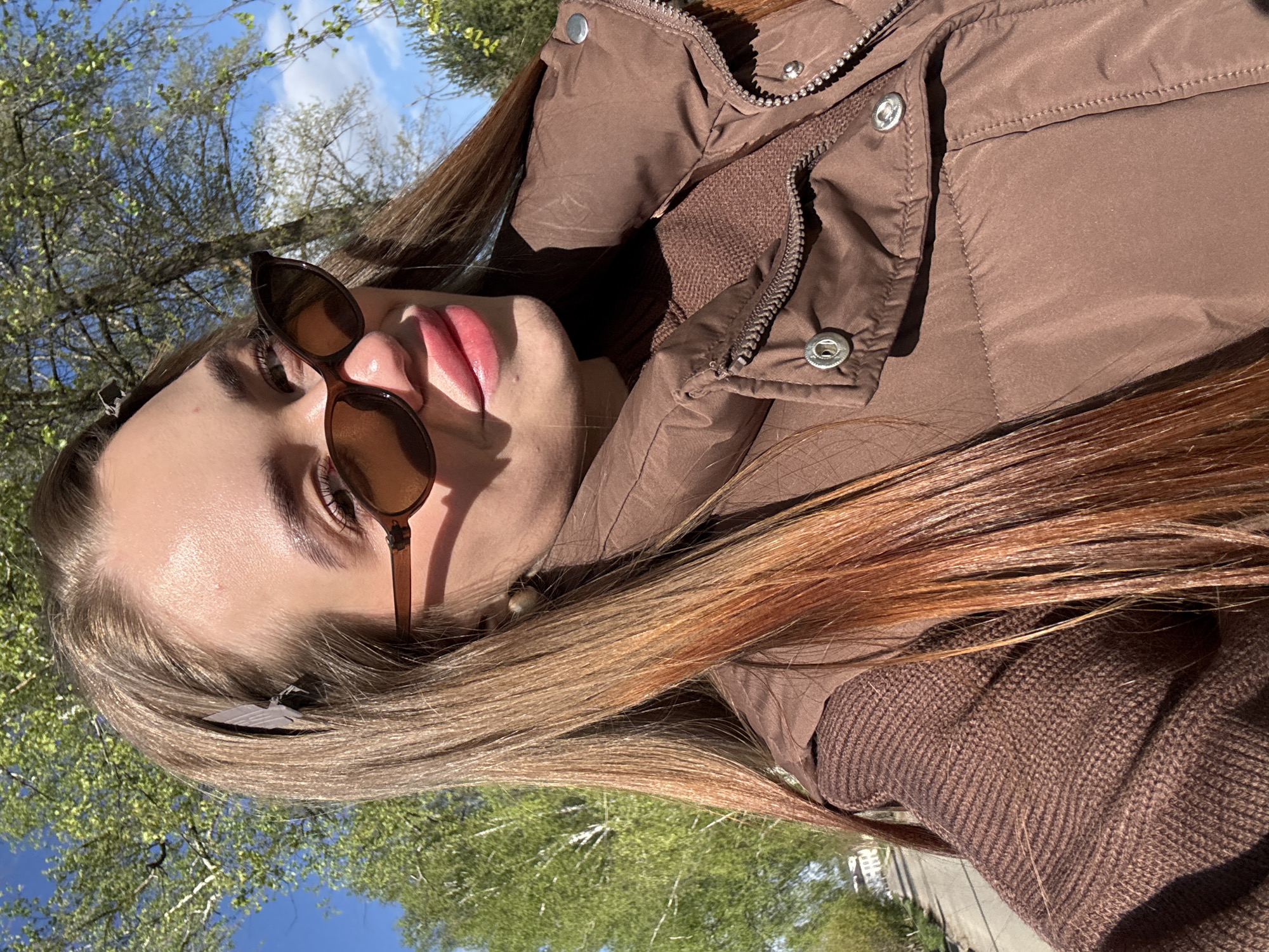 A young woman is the primary subject of this low-angle, outdoor photograph. She is wearing brown sunglasses and a brown puffer jacket. Her long hair has highlights. The background shows a clear blue sky with tree branches and some greenery, suggesting a park-like setting. The location is identified as Chaikovsky, Russia, based on the prompt.