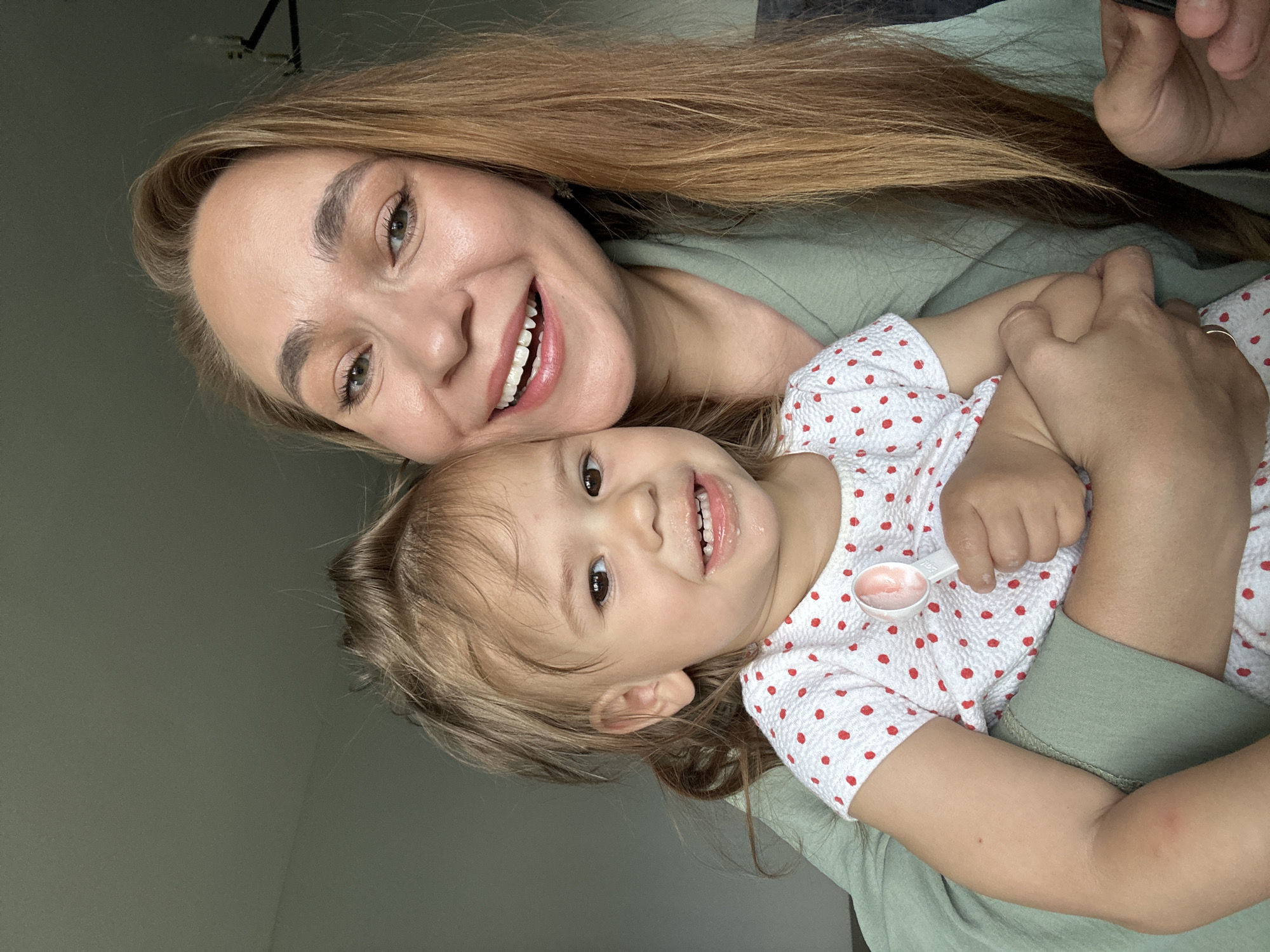The media file is a close-up, low-angle selfie of a woman holding a young child. The woman, with long blonde hair, is smiling broadly, showing her teeth. She is wearing a green top. The child, facing the camera, also has a wide smile and is wearing a white top with red polka dots. The child's right hand is visible, holding a small white measuring spoon with a pink substance, possibly medicine or food. They are positioned closely together, suggesting an affectionate moment. The background is a plain, muted green wall, with a hint of a ceiling fixture at the top left corner. The lighting is soft and appears to be natural, likely from a window, suggesting it could be daytime. The overall emotion conveyed is happiness and warmth between the two individuals. There is no visible text in the image. The location context provided is Kharnavy, Russia.