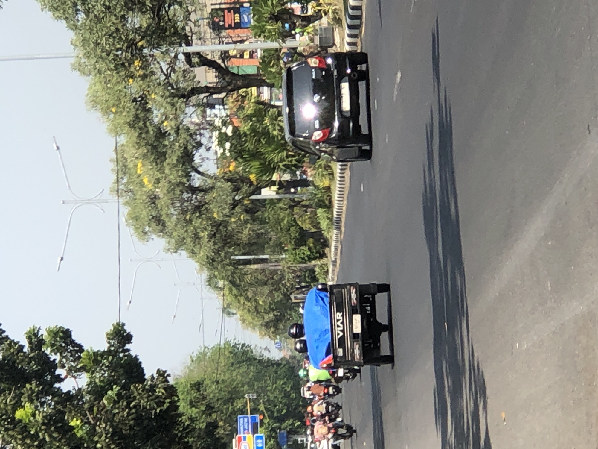 The image shows a street scene in Larangan, Indonesia, on a sunny day. In the foreground, a black car is driving on the road, its headlights reflecting the sun. To the left of the car, a small utility vehicle with a blue tarp covering its cargo area is also driving on the road. The vehicle has "VIAR" written on the back. Several motorcycles with riders are visible in the background, as well as trees and streetlights lining the road. The sky is clear and blue.