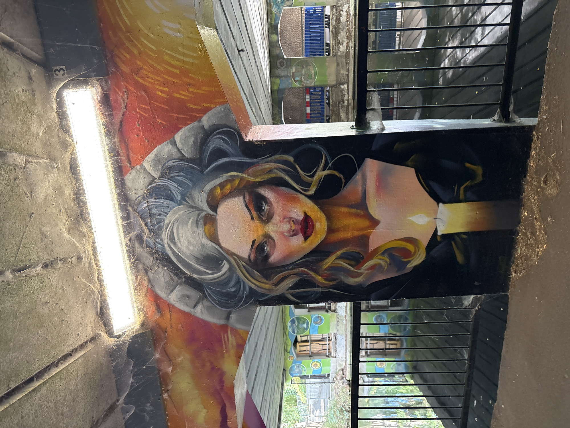 The image shows a mural of a woman's face painted on a wall. She has blonde hair with streaks of grey, dramatic eye makeup, and bright red lipstick. She appears to be looking directly at the viewer. In front of her, a lit candle is painted. The mural is located under a concrete bridge or underpass, as evidenced by the exposed concrete ceiling and supporting pillars. A fluorescent light fixture hangs from the ceiling, illuminating the mural. There are also cobwebs visible on the ceiling and around the light fixture. The background shows a body of water, likely a canal, with a metal railing in the foreground. On the far side of the water, there are some structures with colorful circular designs and wooden elements. The mural is in a public area, as indicated by the presence of a railing and the overall urban setting. The number "3" is visible on the concrete ceiling to the right of the light fixture. The mural appears to be located on a wall near the River Chelmer in Chelmsford, United Kingdom, likely under one of the bridges that cross it.