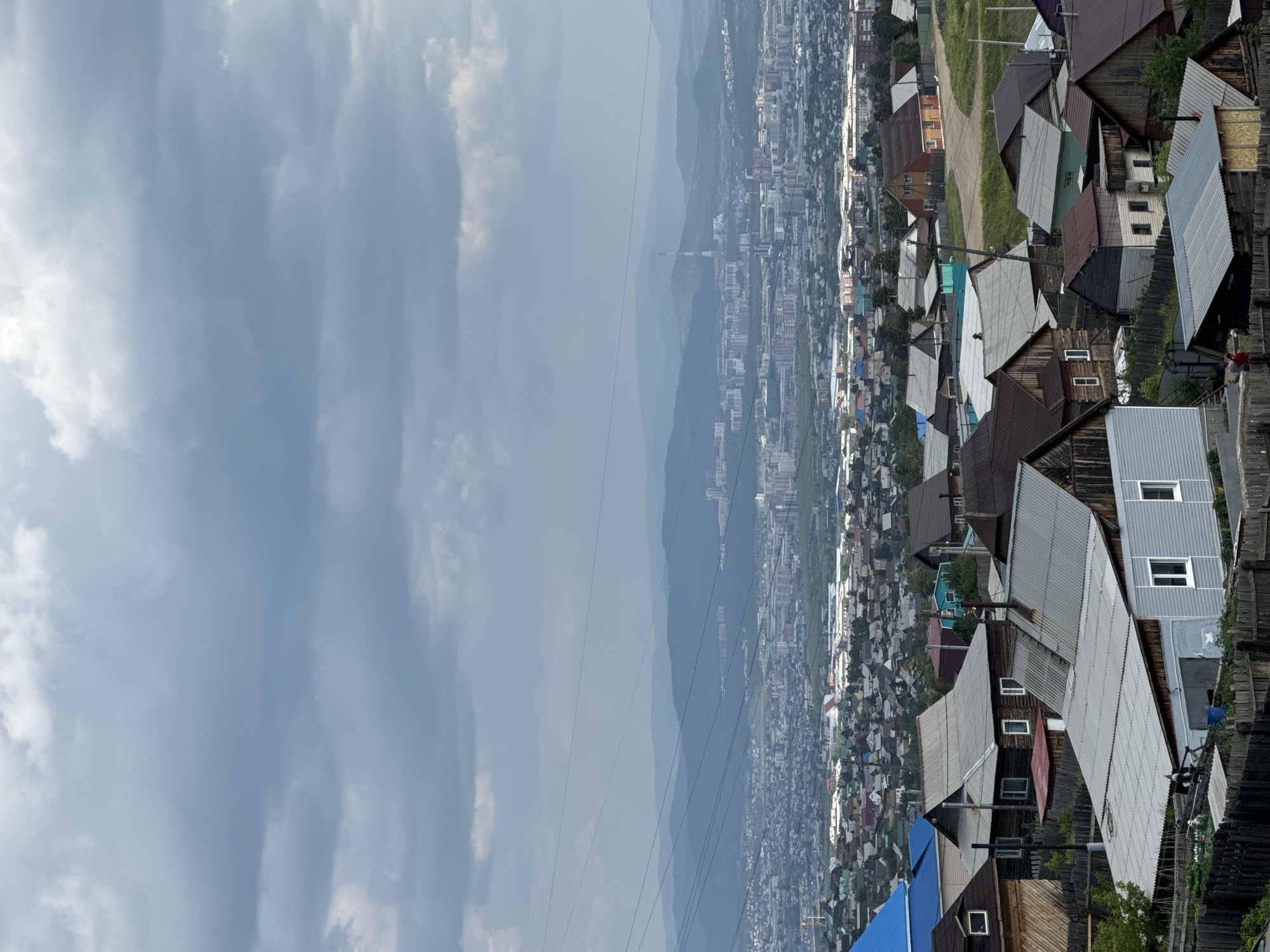 The image depicts a panoramic view of the outskirts of Ulan-Ude, Russia, under a cloudy sky. In the foreground, a collection of modest residential structures, primarily single-story houses with corrugated metal roofs, are clustered together. These homes exhibit varied architectural styles and materials, including wooden siding and fences, suggesting a developing or rural-urban fringe area. Some houses have visible windows and chimneys, and a few appear to be under construction or in disrepair.
Beyond the immediate residential area, the cityscape of Ulan-Ude unfolds, characterized by a dense arrangement of buildings, including multi-story apartment blocks and industrial structures. The city stretches towards a backdrop of rolling, forested hills that fade into the atmospheric haze. A tall, slender industrial smokestack or tower is prominent on the horizon. Numerous power lines crisscross the mid-ground of the image, indicating the presence of electrical infrastructure. The overall scene conveys a sense of a large, inhabited area transitioning from lower-density housing to a more urbanized center, set against a natural, mountainous landscape.