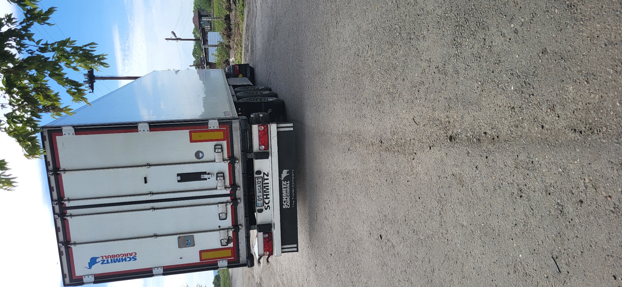 An outdoor, eye-level, vertical, medium shot shows the rear of a white Schmitz Cargobull semi-trailer truck parked on a paved surface that appears to be a road or industrial area. The truck has a large white trailer with red and white trim and multiple latches on the rear doors. The Schmitz Cargobull logo, featuring a blue elephant, is visible on the left side of the trailer. A license plate with the characters "01 AGA 01" is affixed to the rear bumper. Below the license plate, a black bumper displays the text "SCHMITZ CARGOBULL The Trailer Company." with a smaller elephant logo. The truck's tires and part of its undercarriage are visible. The environment includes a clear blue sky with scattered white clouds, green foliage from a tree in the upper right corner, and some distant buildings and utility poles in the background, suggesting an urban or semi-urban setting. The ground surface is asphalt with visible aggregate. No people are present in the image.