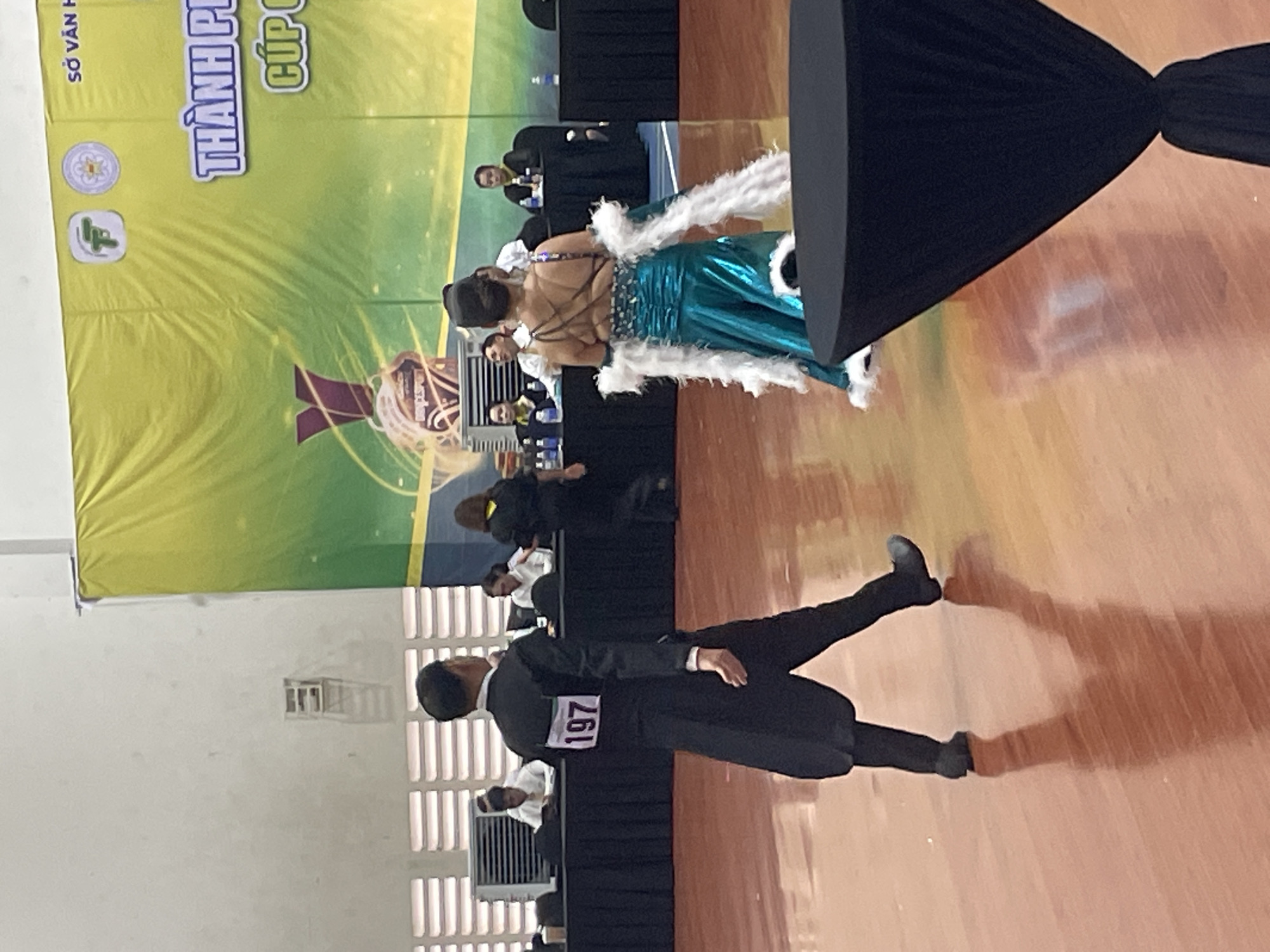 The image shows a ballroom dancing competition or performance at an indoor venue. In the foreground, a male dancer in a black tailcoat with a white bib displaying the number "197" is walking away from the camera. To his right, a female dancer in a shimmering turquoise gown with white feather accents on her arms is also walking away from the camera. In the background, a large green and yellow banner is visible with Vietnamese text and logos. Several people are seated at tables behind the dancers, possibly judges or spectators. The floor is a polished wooden surface. This event is likely taking place in Thuận An, Vietnam, as indicated by the banner which appears to be from the Department of Culture and Information of Thuận An City.