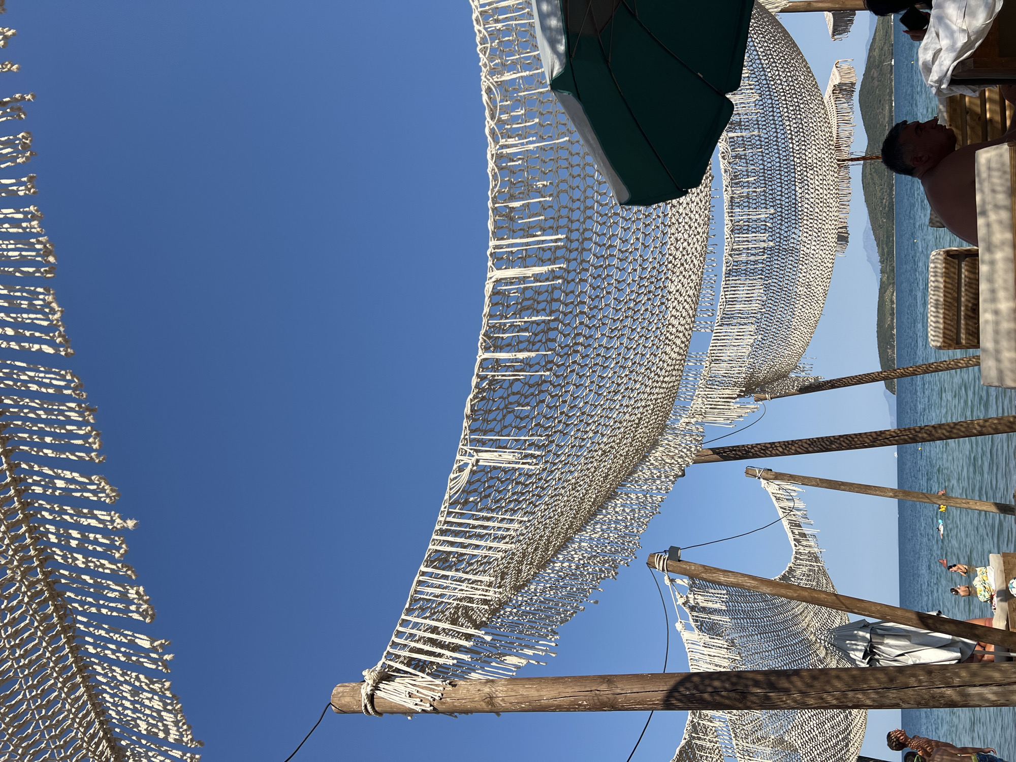 A view of the beach at Nea Peramos, Greece, on a sunny day. The sky is a clear, vibrant blue, with a few scattered, indistinct clouds visible in the distance. In the foreground, several wooden poles support a woven netting canopy with a decorative fringe of hanging ropes. The netting creates a dappled shade effect.
To the right, a green beach umbrella is partially visible, angled upwards. The sea is calm and blue, stretching to the horizon. Several people are in the water, some swimming and others relaxing. In the mid-ground, a man is seated on a wooden lounge chair, facing towards the sea. His gaze is directed downwards.
The beach appears to be part of a resort or a beach bar, with more lounge chairs and umbrellas visible further back. The backdrop features rolling green hills and islands. The overall impression is one of a tranquil summer day at a coastal destination.
