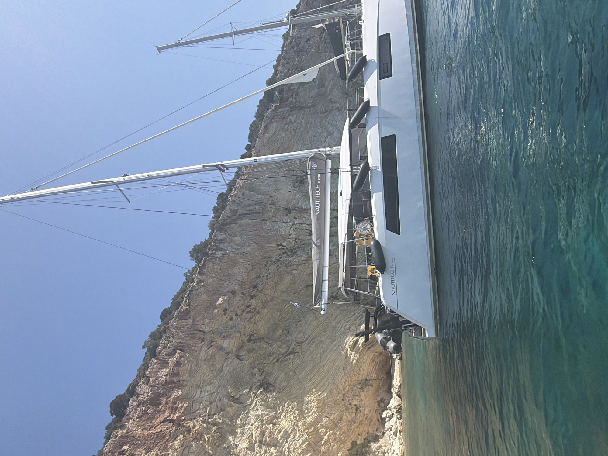 The image shows a white catamaran sailing boat docked near a rocky cliff face. The water is clear and turquoise, with some ripples on the surface. The cliff is tall and imposing, with layers of rock visible. The sky is a clear blue. The boat has two masts and sails. The lettering "NAUTITECH 44 OPEN" is visible on the hull of the boat. A Greek flag is flying from a rope attached to the boat. The scene appears to be in a secluded cove or bay. The location is the Municipal Unit of Kastos, Greece.