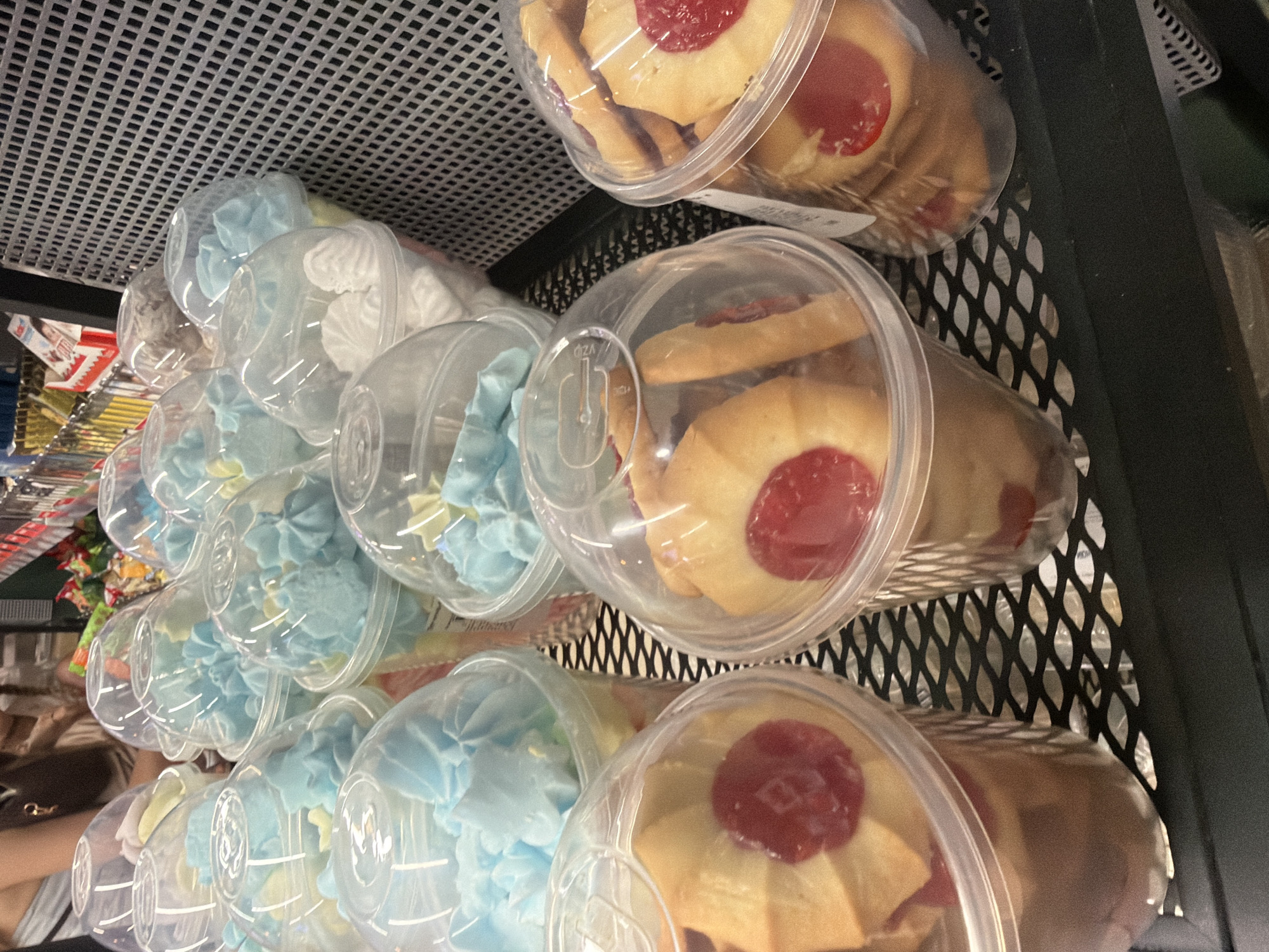 In an indoor market stall, a display of sweets is presented. The foreground features several plastic cups filled with jam cookies. These cookies are round and golden brown with a red jam filling in the center. Behind the cookies, more plastic cups are visible, some containing light blue meringue swirls and others filled with white meringue. The display is set against a black metal grate background, and a hint of a person is visible in the upper left corner.