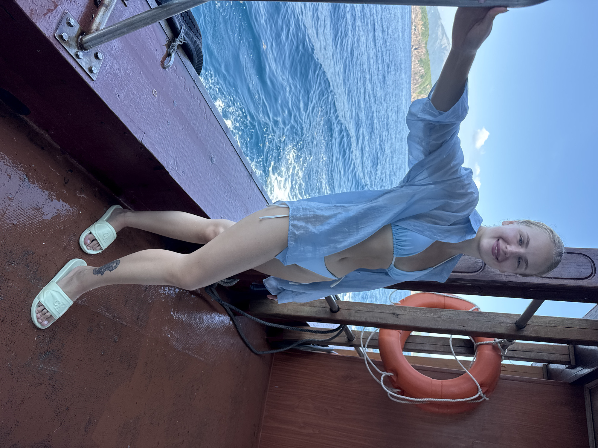 A young woman with blonde hair and a bright smile is enjoying a boat ride on the ocean. She is wearing a light blue bikini and an open light blue button-up shirt, with white slides on her feet. The boat appears to be wooden, with a reddish-brown deck and railing. A bright orange life preserver with white rope is visible, attached to the boat's structure. The woman is leaning on the railing, looking at the camera. Behind her, the deep blue ocean stretches to the horizon, with whitecaps indicating movement and a gentle breeze. In the distance, a lush green, mountainous coastline is visible under a clear, sunny sky. The overall impression is one of a relaxed and joyful vacation experience. The image suggests it is daytime, with ample sunlight contributing to the vibrant colors of the scene.