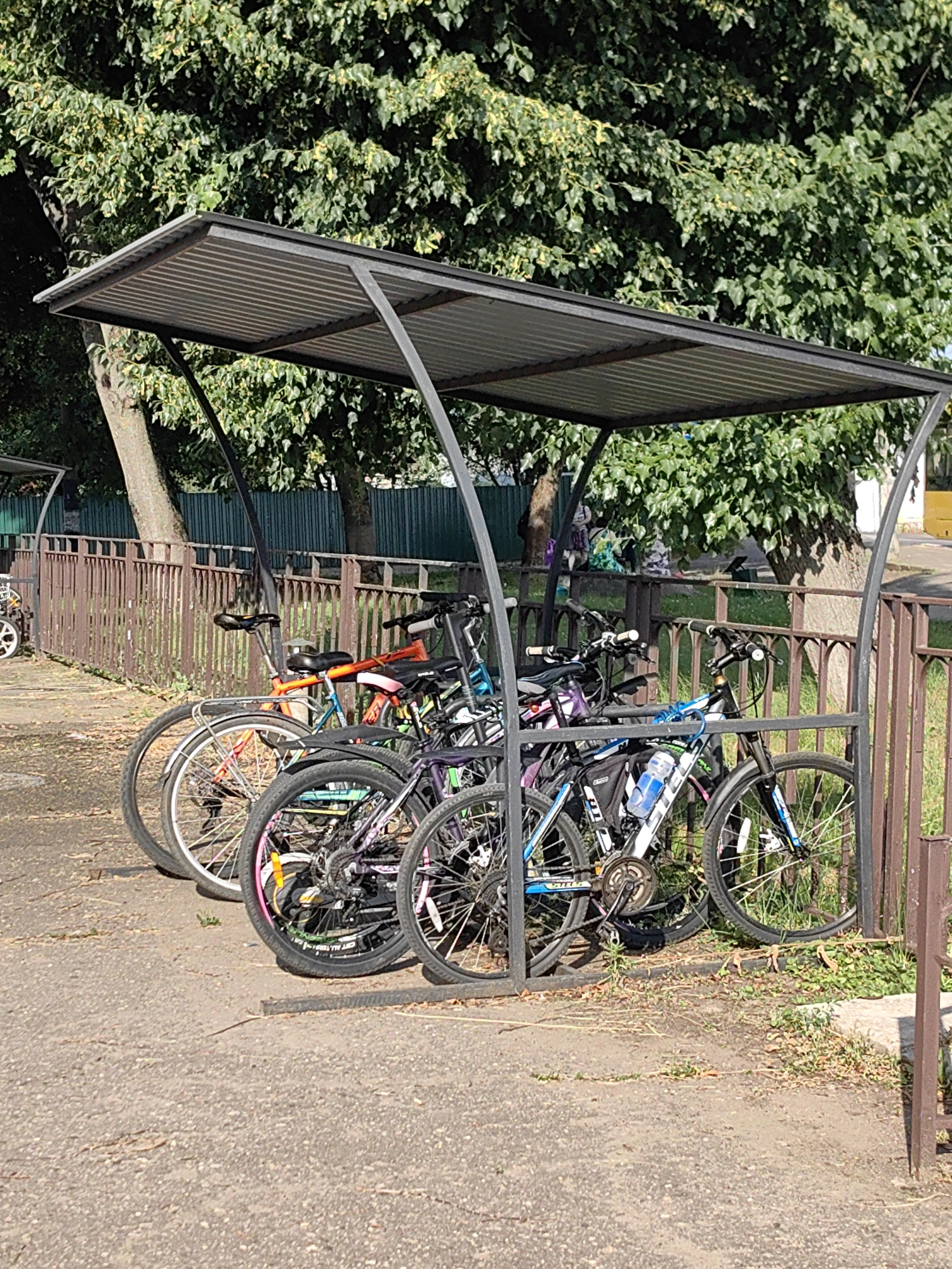 This is a vertical, medium-shot photograph taken on a sunny day in Fanipal, Belarus. The image depicts a row of bicycles parked under a metal canopy designed to shelter them from the elements. The canopy has a corrugated metal roof and dark grey metal support posts.
The bicycles are of various colors and designs, including a bright orange and blue bike, a purple and black bike, and a black and blue bike with a water bottle mounted on the frame. Some of the bicycles have colorful accents on their rims. They are parked closely together, suggesting a communal bike rack.
In the background, a rusty brown metal fence runs horizontally across the frame. Beyond the fence, lush green trees with dense foliage dominate the upper portion of the image, casting dappled shadows. The ground beneath the canopy and in front of the fence is a mix of asphalt and dry, patchy grass with fallen leaves. The overall scene appears to be an outdoor area, likely in a residential or public space, designed for bicycle parking. The bright sunlight indicates it is daytime. There is no visible text in the image.