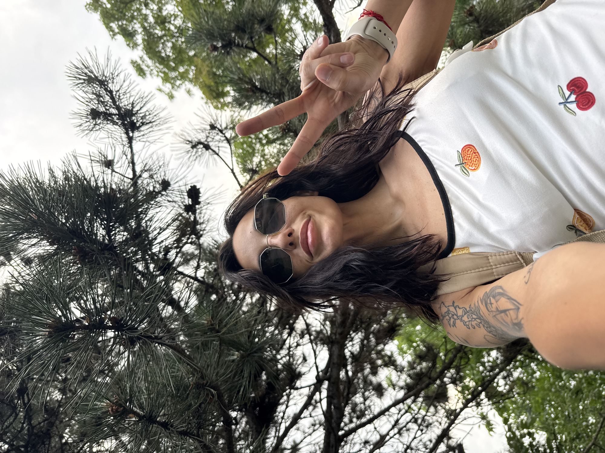 A woman with dark, shoulder-length hair is captured in a low-angle selfie, taken in the city of Pleven, Bulgaria. She extends her left hand forward, forming a peace sign with her index and middle fingers. She wears dark, octagonal-framed sunglasses, obscuring her eyes, and her lips are slightly parted in a subtle smile. Her attire consists of a white sleeveless top with black trim, featuring embroidered fruit designs including an orange, a cherry, and a partial lemon. A beige backpack strap is visible over her right shoulder. On her left wrist, she wears a light-colored smartwatch and a red string bracelet. Her left upper arm displays multiple tattoos, including a vine-like pattern and an abstract, possibly heart-shaped, motif. The background is dominated by the dense, dark green foliage of a conifer tree, likely a pine, with visible needles and small pinecones, positioned primarily above and to the left of the woman. To the upper right, lighter green deciduous tree foliage is visible against an overcast or bright sky. The image is taken from a low vantage point, looking upwards towards the subject and the overhead tree canopy.