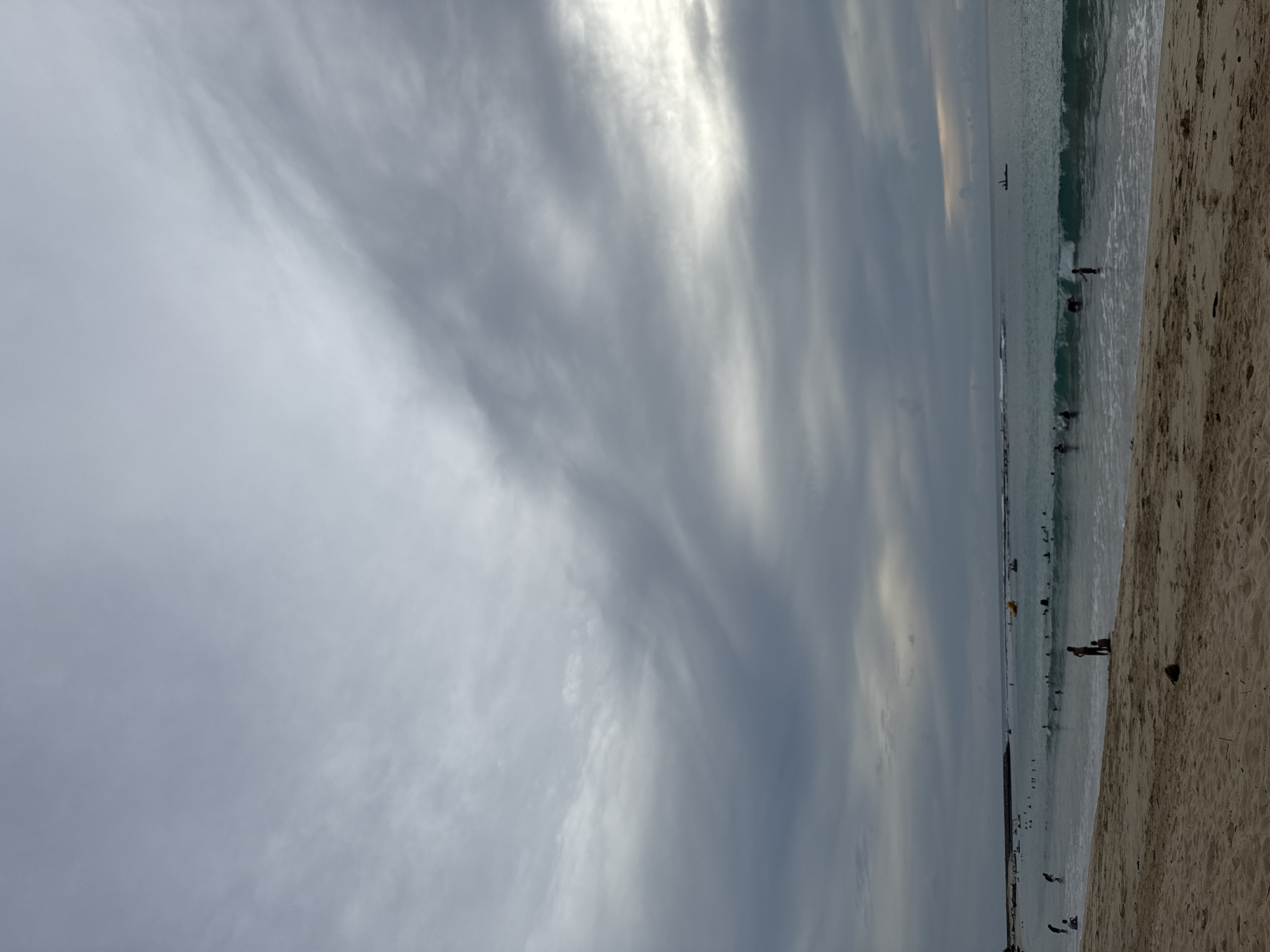The image depicts a beach scene at what appears to be Meunasahbale, Indonesia. The sky is overcast with dramatic grey clouds, with a sliver of brighter sky visible on the right horizon. The ocean is a turquoise color with moderate waves, and several people are visible in the water, some appearing to be surfing or playing. There is a yellow inflatable object in the water, and a small dark boat is further out at sea. On the sandy beach in the foreground, a few individuals are present, and some debris, possibly a coconut, is on the sand. The overall impression is a somewhat moody, but active, beach day.
