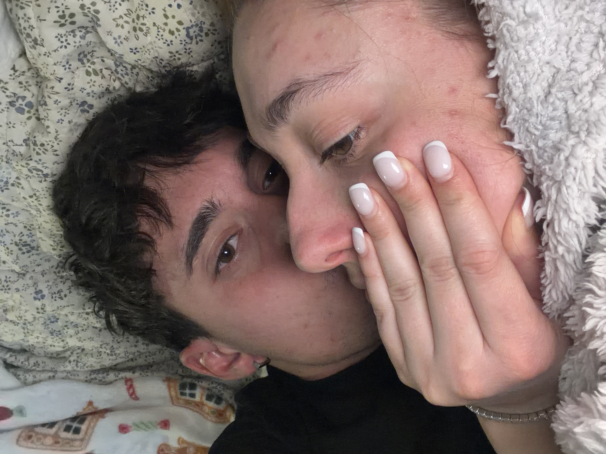 The image captures an intimate and tender moment between two individuals, likely a couple, lying very close together indoors, possibly in a bedroom setting in Sofia, Bulgaria. The perspective is a close-up, looking down on the subjects.
On the left, a person with dark, curly hair, who appears to be male, has their head resting against the side of the other person's face. Their brown eye is visible, looking towards the camera with a soft gaze. They are wearing a black top.
The second individual, likely female, is partially obscured by the first person's head. Her right hand, featuring a classic French manicure with white tips, is gently placed against her own cheek or the first person's face. Her brown eye is cast downwards or slightly away, contributing to a contemplative or relaxed expression. Her skin shows some minor blemishes, indicating a natural appearance. A fluffy, white blanket or robe is draped around her right side, and a silver-colored beaded bracelet is visible on her wrist.
The background includes a patterned quilt or comforter with small blue and green floral designs on a light fabric, and another blanket or sheet featuring a festive pattern with gingerbread houses and candies. The soft lighting suggests an indoor setting, likely in the morning or evening. The overall mood is one of closeness, comfort, and affection. No visible text is present in the image.