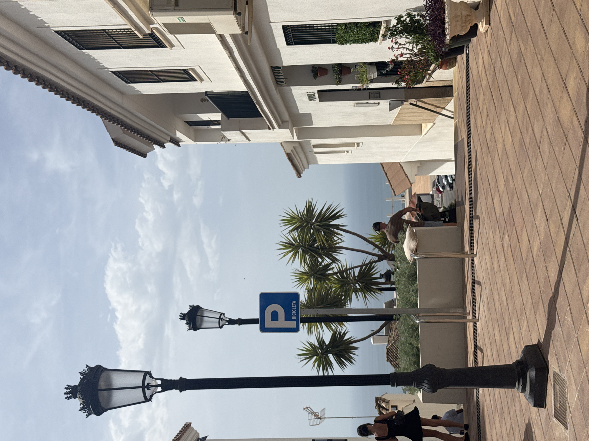 This is an outdoor, eye-level, medium shot of a street scene in Benalmádena, Spain. The image is captured on a sunny day with a partly cloudy sky. The foreground is dominated by a textured reddish-brown tiled pathway, casting a long shadow from a tall, black, ornate lamppost on the left.
In the mid-ground, a white building with white stucco walls and a red-tiled roof stands on the right, featuring barred windows and balconies. Next to it, a pathway descends, with another building on the left that is partially visible. Between the buildings, a few palm trees are seen, along with some shrubbery.
On the left side of the pathway, a woman in a black dress walks away from the camera. Further back, another person, possibly a child, is seated. In the center of the frame, a blue parking sign for bicycles is mounted on a pole next to a lamppost. A man is pushing a cart on the pathway behind the sign.
In the background, the blue sea is visible, with some rooftops and other buildings interspersed. The overall scene depicts a charming street in a Spanish coastal town.
