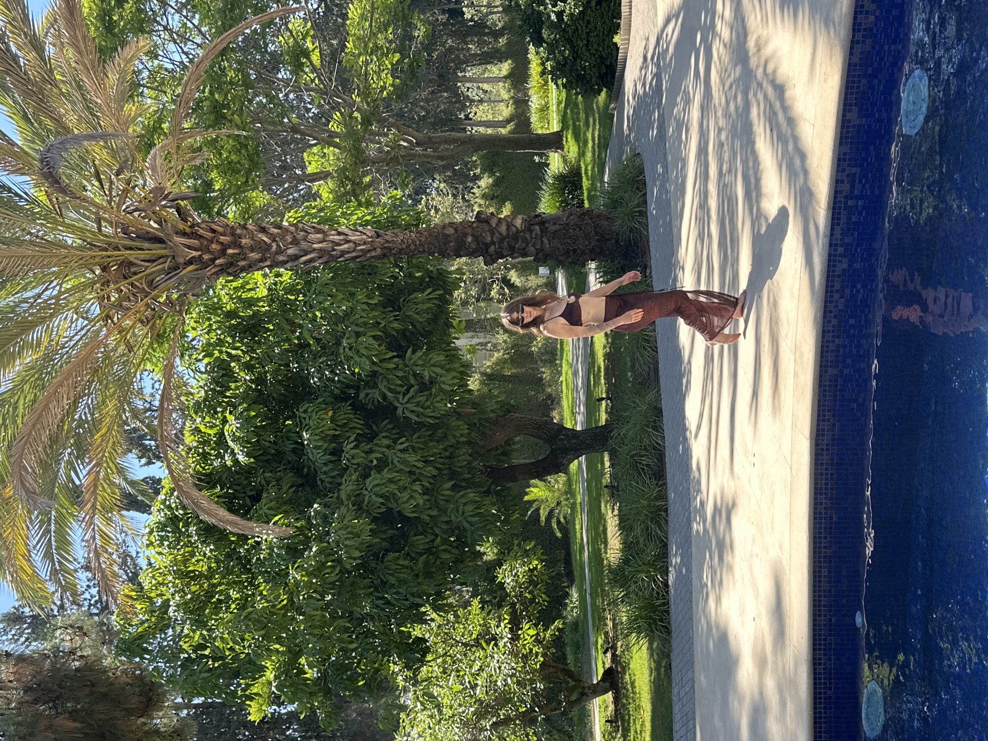 The image depicts a woman wearing a brown bikini and a sheer brown skirt walking barefoot on a paved area beside a swimming pool. She is positioned in the middle ground, with a large palm tree to her right and lush green trees and foliage in the background. The sun casts dappled shadows on the ground from the palm fronds. The swimming pool, with its dark blue tiled interior, is in the foreground, partially reflecting the surroundings. The setting appears to be an outdoor residential area, possibly a backyard or garden, consistent with the affluent character of Savyon, Israel, known for its luxurious villas and extensive gardens. The overall atmosphere is one of leisure and warm weather.