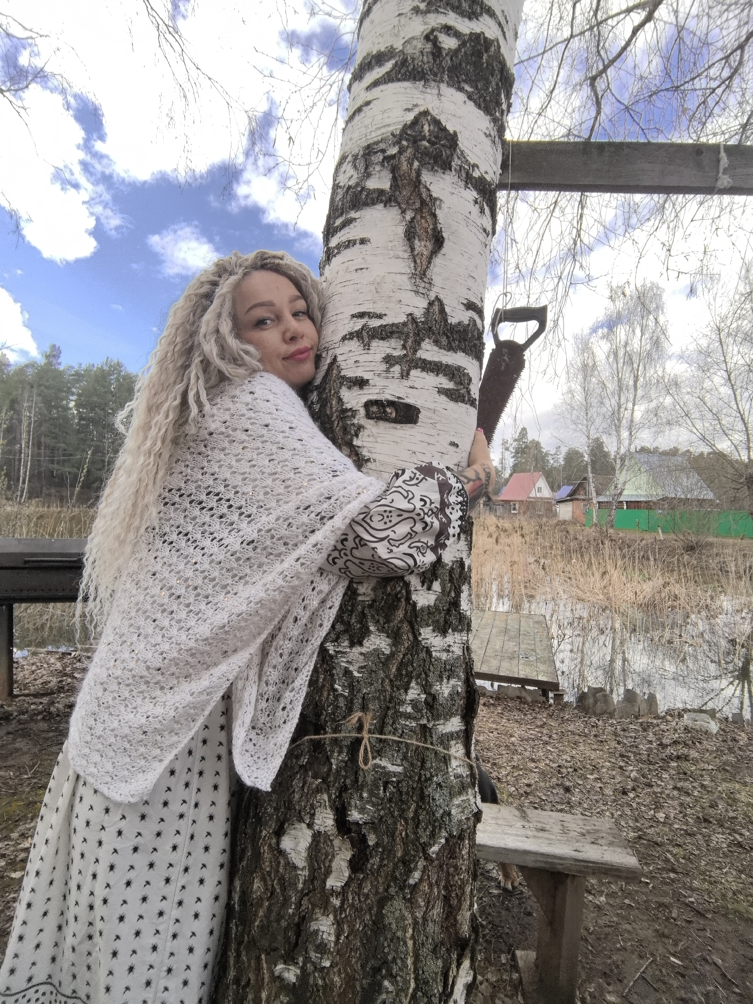 A fair-skinned woman with long, curly, blonde hair hugs a birch tree. She wears a white dress with small black starburst patterns and a white, lacy shawl. A handsaw hangs from a branch on the tree. In the background is a body of water with reeds, wooden docks, and houses with colorful roofs. The houses appear to be in a rural area, with trees, grass, and leaves on the ground. The sky is blue with scattered clouds. This location is Noviy, Russia.
