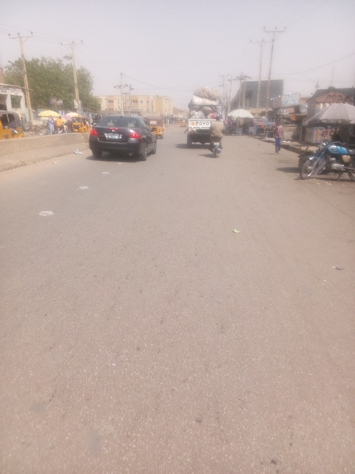 The image displays a daytime street scene in Dakata, Nigeria, characterized by a hazy atmospheric condition that softens distant features. The central element is a granular, light brown-grey asphalt road extending into the background, appearing to be a multi-lane thoroughfare. Small, scattered white debris is visible on the road surface in the foreground and midground.
In the midground, traffic proceeds away from the viewer. A dark-colored Honda Civic sedan with a rear license plate (partially legible as "YBC 9XX") occupies the left lane. In the right lane, a white Toyota pickup truck, heavily laden with large, irregularly shaped bundles wrapped in light-colored material, is visible. A person on a small motorcycle follows directly behind the pickup truck.
To the left of the road, a concrete median separates the main carriageway from a roadside area. This area contains multiple yellow motorized rickshaws/tricycles and several pedestrians. Beyond this, white-walled buildings are visible, interspersed with green foliage from trees, and utility poles supporting power lines extend along the street.
To the right of the road, a series of market stalls or shops line the thoroughfare, featuring various coverings including white and striped umbrellas or canopies. Several individuals are present in this area, some walking. A light blue motorcycle is parked on its kickstand on the right shoulder. Similar to the left side, utility poles with power lines and additional buildings are discernible in the background. The scene depicts a busy urban commercial environment.