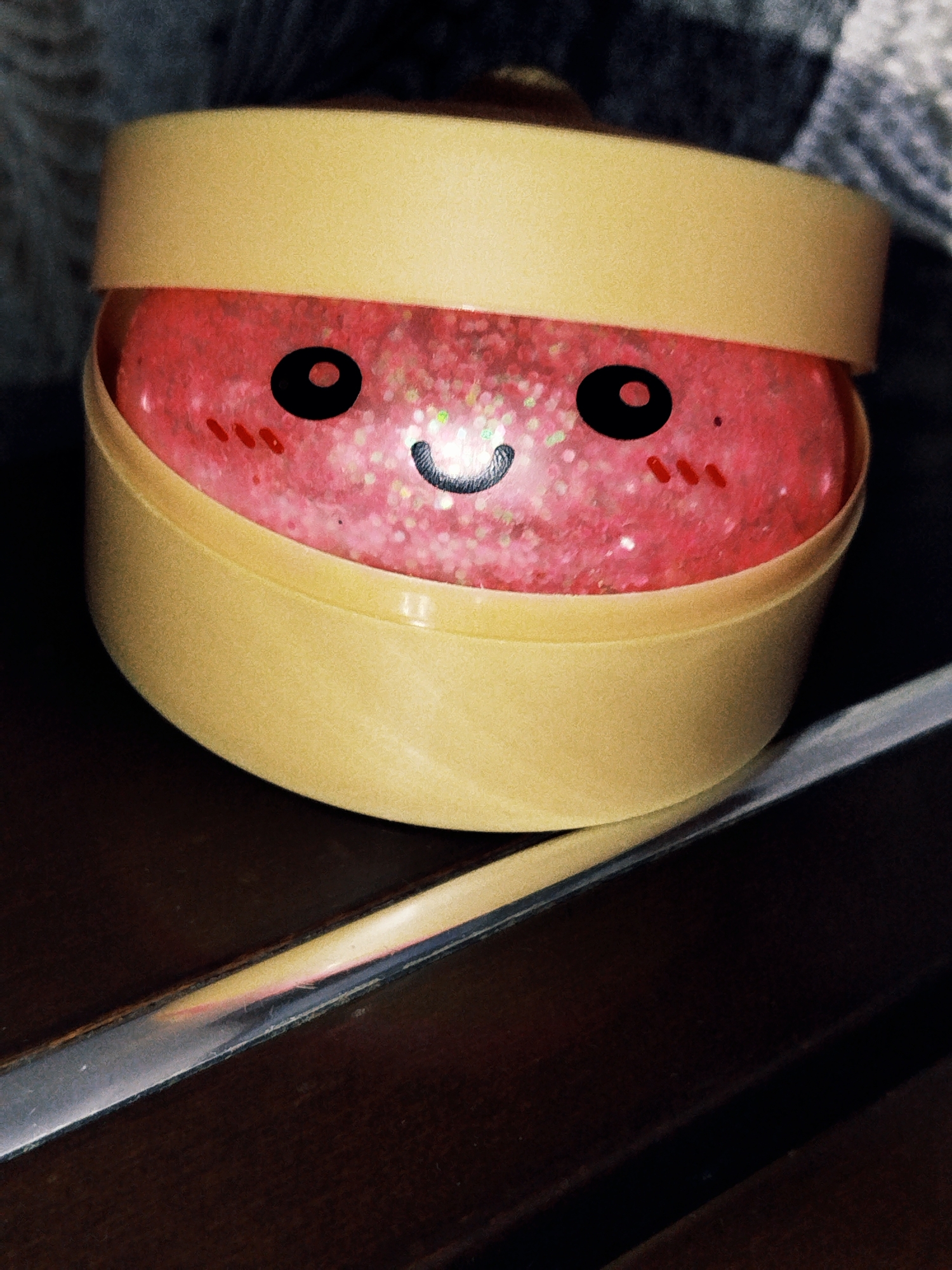 The image shows a close-up of a pink, glittery substance inside a light yellow container. The substance has a cartoon face drawn on it, with black circles for eyes, a smiling mouth, and red dashes for blushing cheeks. The container appears to be a jar or tub with a lid that is partially open. The background is dark and out of focus, with a hint of a grey and black textured fabric. A reflective surface, possibly a table, is visible in the foreground.