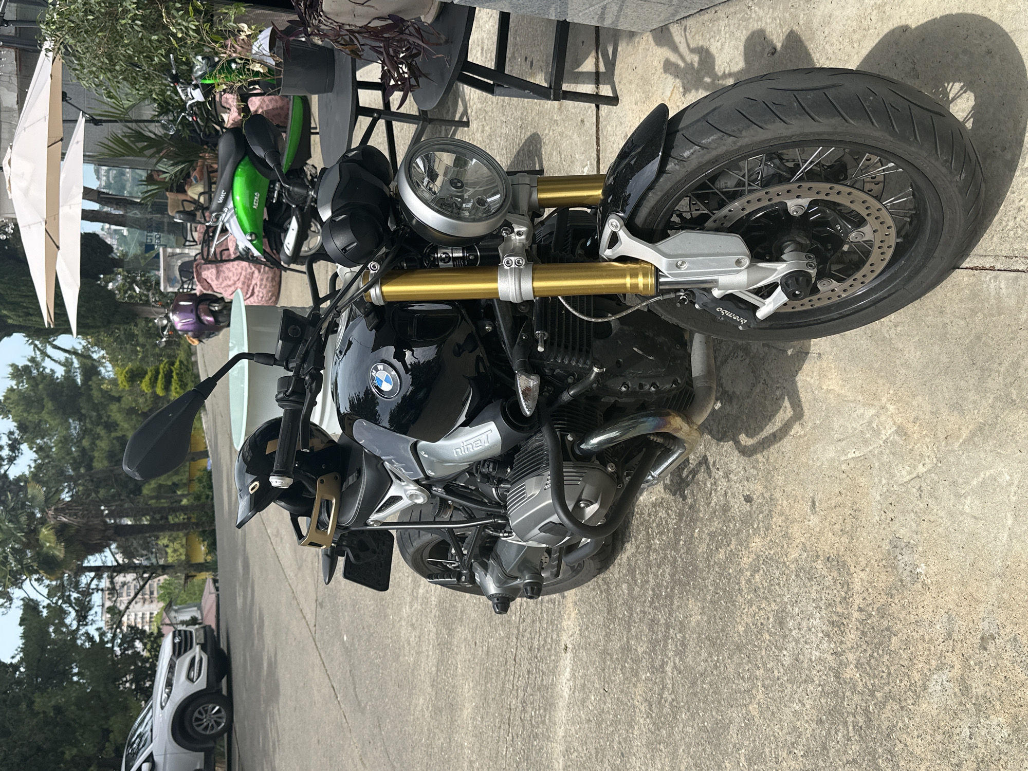 A black BMW R nineT motorcycle is parked on a concrete surface near a cafe in Sochi, Russia. The motorcycle is the primary subject and is viewed from the front and slightly to the right. It has gold-colored front forks, a round chrome headlight, and a black seat. A black helmet with a gold trim rests on the handlebars. In the background, a white SUV is parked to the left. Further back, several scooters are visible, along with outdoor cafe seating, umbrellas, potted plants, and buildings. The scene is brightly lit by sunlight, casting shadows on the ground. The environment suggests a warm, sunny day in a city setting, likely a tourist area given the presence of cafes and various vehicles.