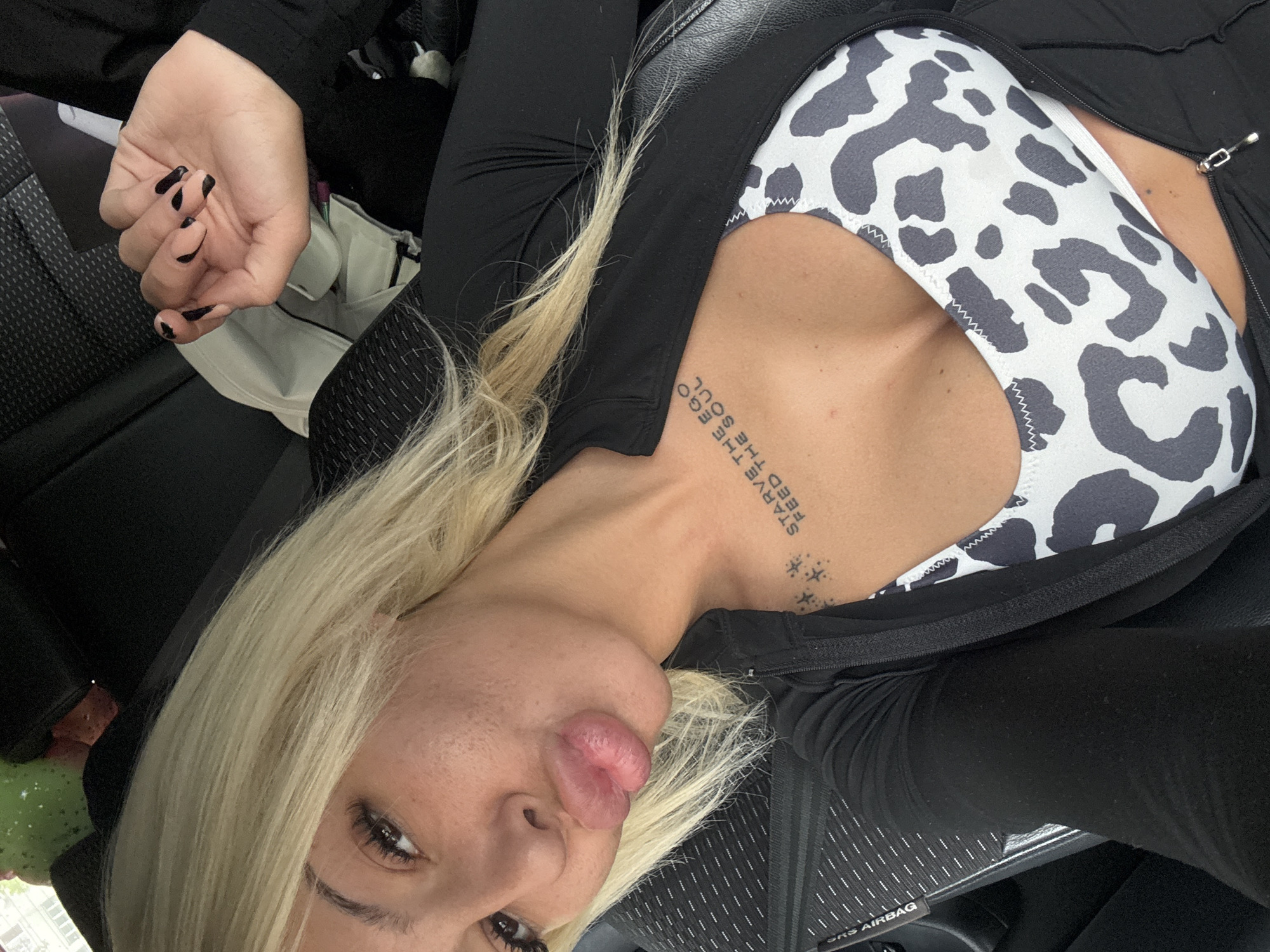 A blonde woman is depicted in a close-up, high-angle selfie, seemingly lying down or leaning back. She is wearing a black long-sleeved top or jacket, partially unzipped, revealing a white and black patterned bra or crop top underneath, resembling a cow print. Her left hand is visible, featuring black nail polish.
A prominent tattoo is visible on her upper chest/collarbone area, reading "STARVE THE EGO FEED THE SOUL" in uppercase letters, with several small star-like symbols tattooed below the text. Her lips are slightly pouted, and she has dark eye makeup.
The setting appears to be indoors, likely the interior of a vehicle, suggested by the dark, possibly leather, seating and a partial "SRS AIRBAG" label visible on a panel in the bottom right corner. A green object is partially visible in the bottom left of the frame. The lighting suggests daytime. The overall composition is a casual self-portrait.