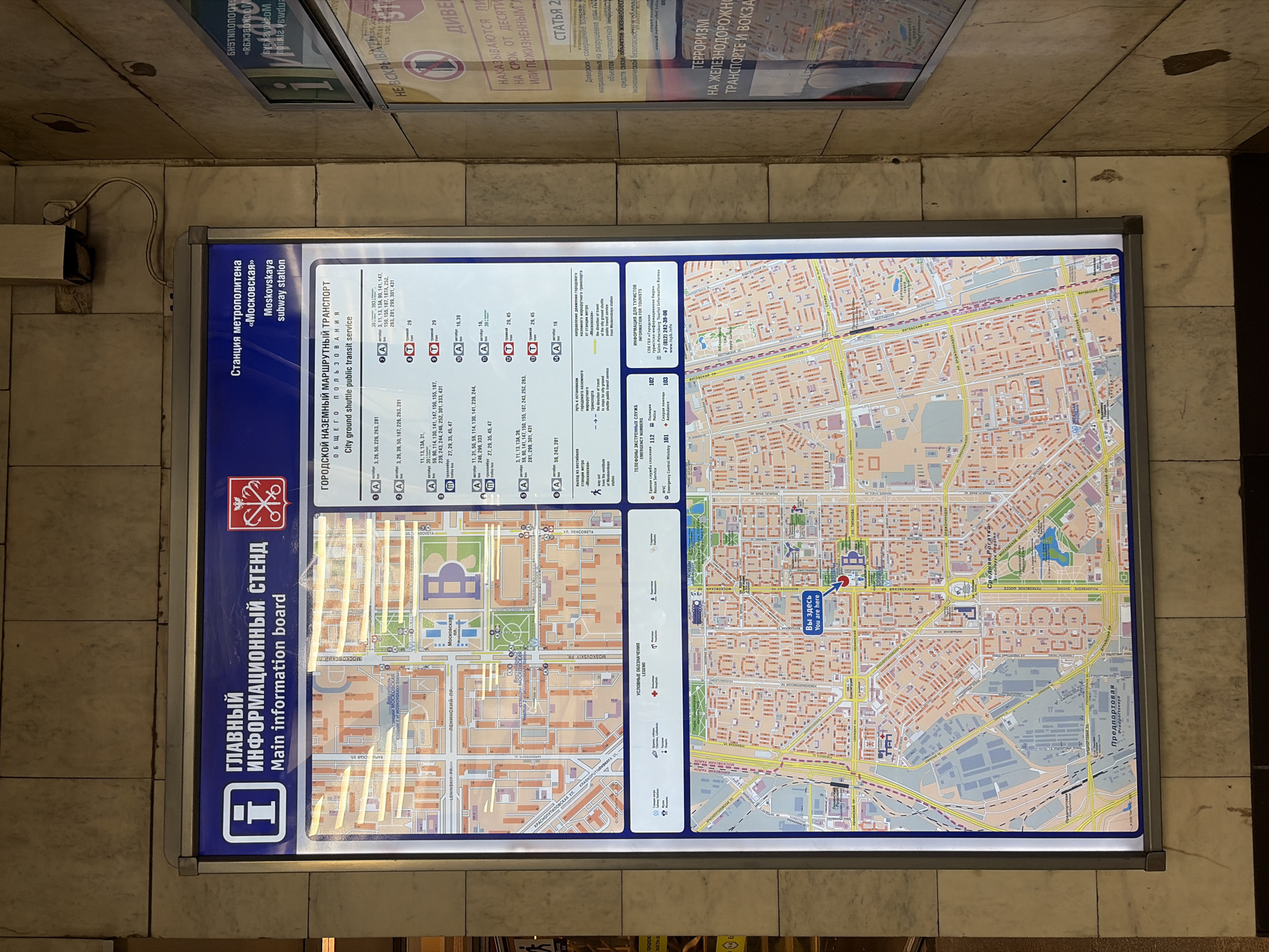 The image shows a "Main information board" inside the Moskovskaya subway station in Saint Petersburg, Russia. The board displays a map of the area around the station, marking the location ("You are here") and showing the routes of city ground shuttle public transit. It includes a legend, emergency numbers, and tourist information. There is also an announcement on the wall about terrorism and sabotage of transportation infrastructure and public facilities.