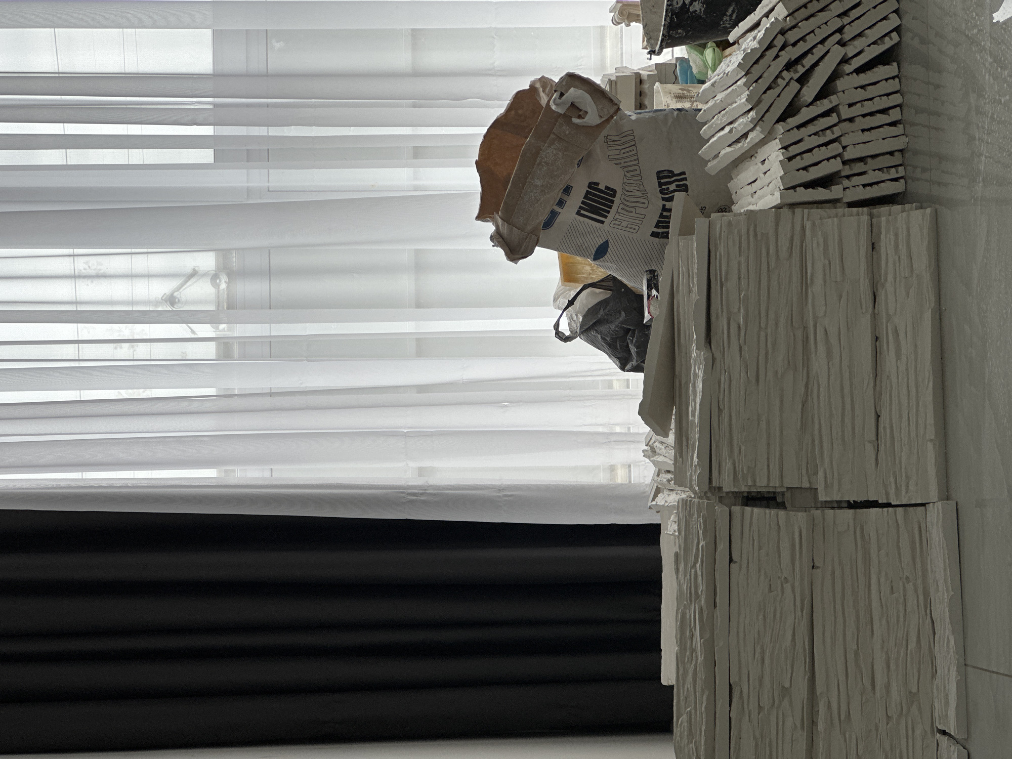 The image captures an indoor scene, likely a room undergoing renovation or construction. On the left, a window is visible, covered by white horizontal blinds and a sheer white curtain, allowing diffused daylight to enter. Below the sheer curtain, a section of a dark-colored, possibly black, curtain or blind is visible.
On the right side of the frame, various construction materials are stacked. A large quantity of white, textured tiles or decorative panels, appearing to be made of gypsum or a similar material, are arranged in piles. Some are stacked vertically, displaying a rough, linear texture, while others are laid horizontally.
Behind these panels, two large bags containing building materials are present. One is a brown paper bag, and the other is a white bag with Russian text printed on it. The visible text includes "ГИПС" (Gypsum), "СТРОИТЕЛЬНЫЙ" (Construction/Building), and "СТР" (an abbreviation, likely related to construction). A crumpled black plastic bag is nestled among the materials. Indistinct items, possibly tools or containers, are also visible in the upper right portion of the image.
No people are depicted, and no specific activity is taking place, but the presence and arrangement of materials strongly suggest an ongoing or paused construction or renovation project. The scene is illuminated by natural light from the window, indicating it is daytime.