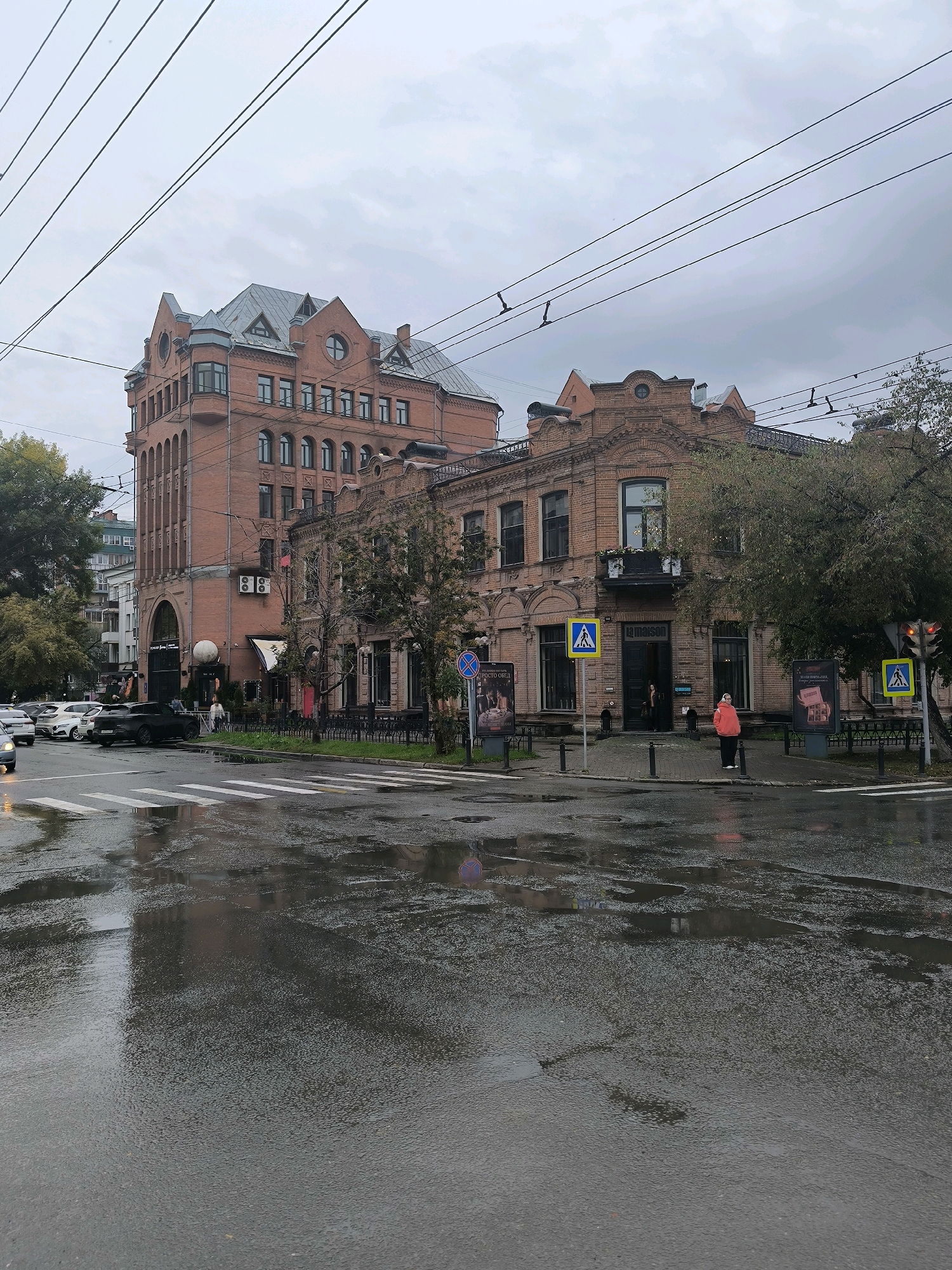 This is a photograph of a street scene in Novosibirsk, Russia, on a cloudy, wet day. The foreground shows a wet asphalt road with reflections of buildings and streetlights. There are white pedestrian crossing lines painted on the road, indicating an intersection. Several cars are visible on the road, including a black SUV.
In the mid-ground, there are two brick buildings. The building on the left is a multi-story structure with a prominent turret and arched windows. The building on the right is a more ornate, older brick building with decorative detailing. There are trees and streetlights lining the sidewalk in front of these buildings. Several pedestrians are present, including a person in a pink jacket standing near the building on the right. Yellow pedestrian crossing signs are visible on poles.
The background shows more buildings and trees, as well as overhead power lines and a cloudy, overcast sky. The overall atmosphere of the image is subdued due to the weather conditions. The scene captures a typical urban environment in Russia during inclement weather.