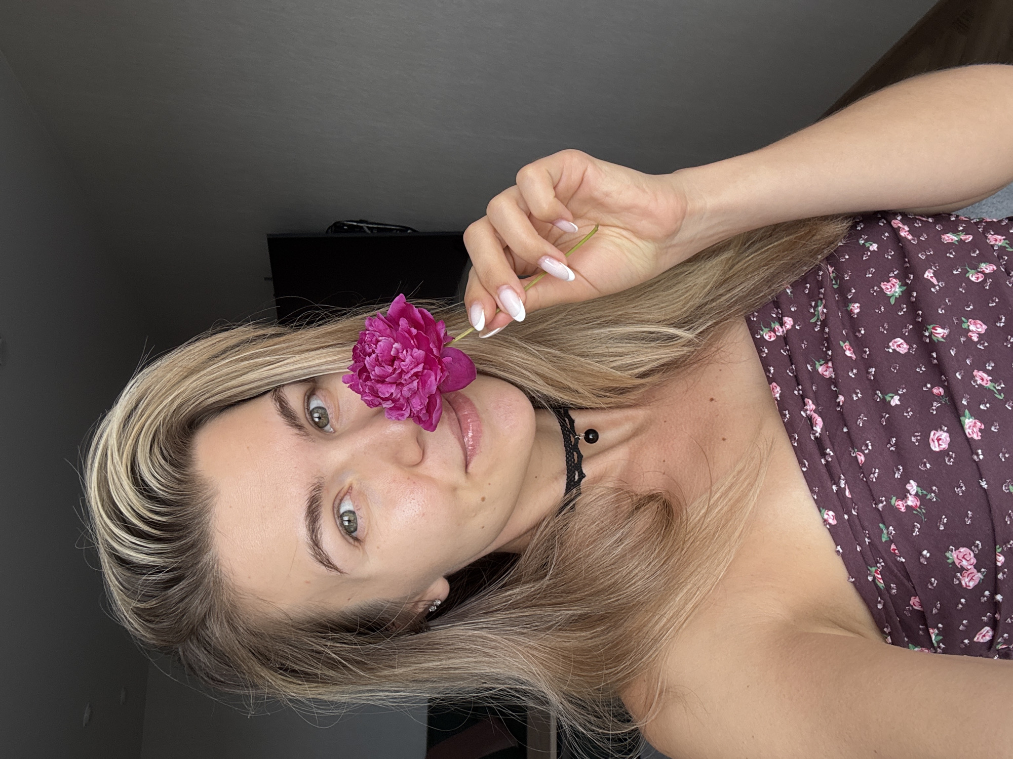 A young Caucasian woman with blonde hair and green eyes is shown in a medium close-up shot, holding a vibrant pink flower near her face. She is wearing a strapless, dark-colored top with a floral pattern. The woman has on a black choker necklace with a small black charm. Her nails are manicured with white French tips. The background is a plain, dark gray wall. The lighting suggests an indoor setting, possibly a room in a home.