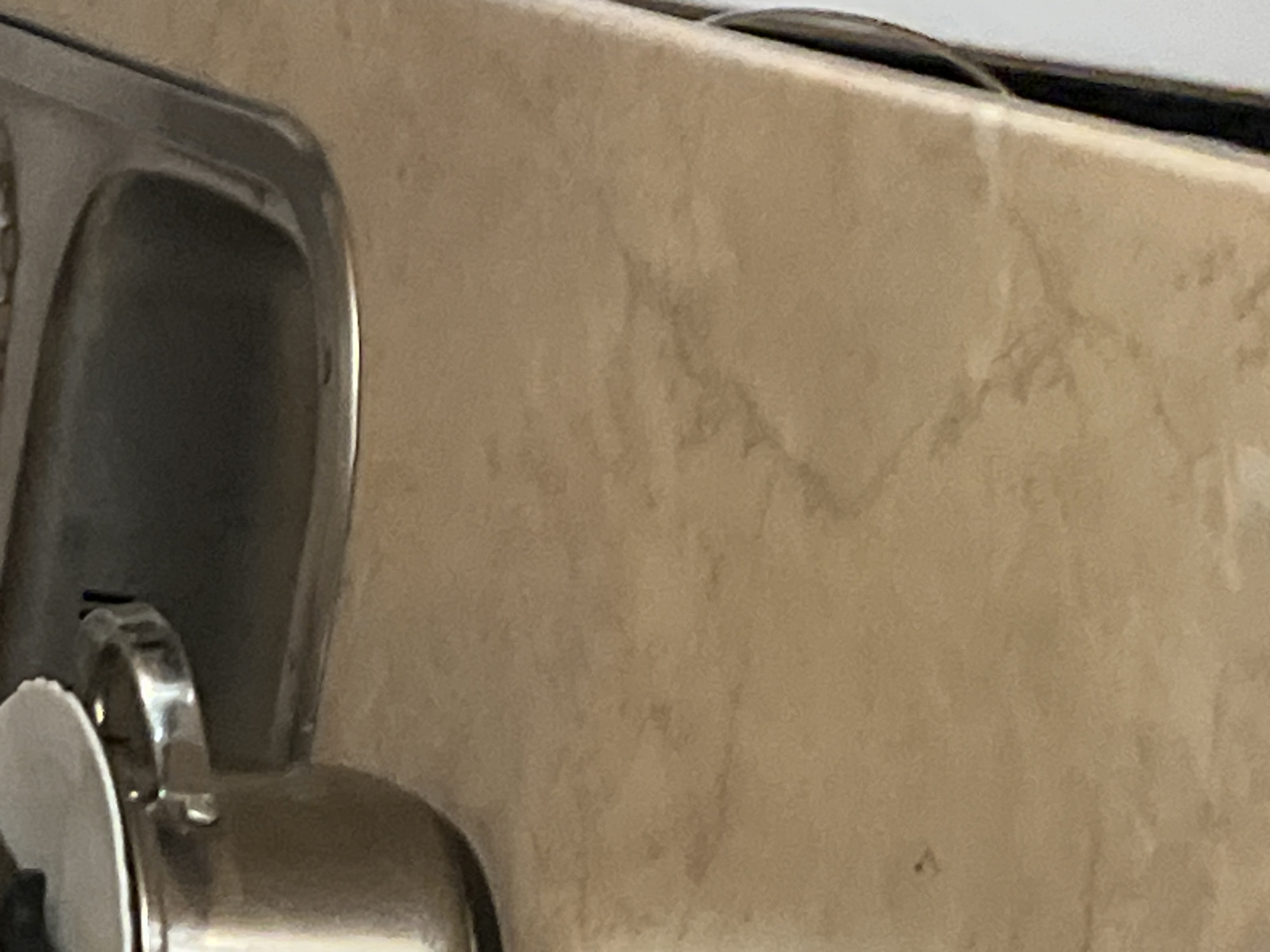 A close-up shot captures a stainless steel pot with a white lid, positioned on a tan countertop. The pot has a handle with a curved rim. To the right of the pot, a stainless steel sink is visible, its interior appearing dark. The countertop itself exhibits a subtle marbled pattern with faint brown streaks. The lighting suggests an indoor environment, possibly a kitchen. The image does not contain any identifiable landmarks or contextual clues to determine its location within a city.
