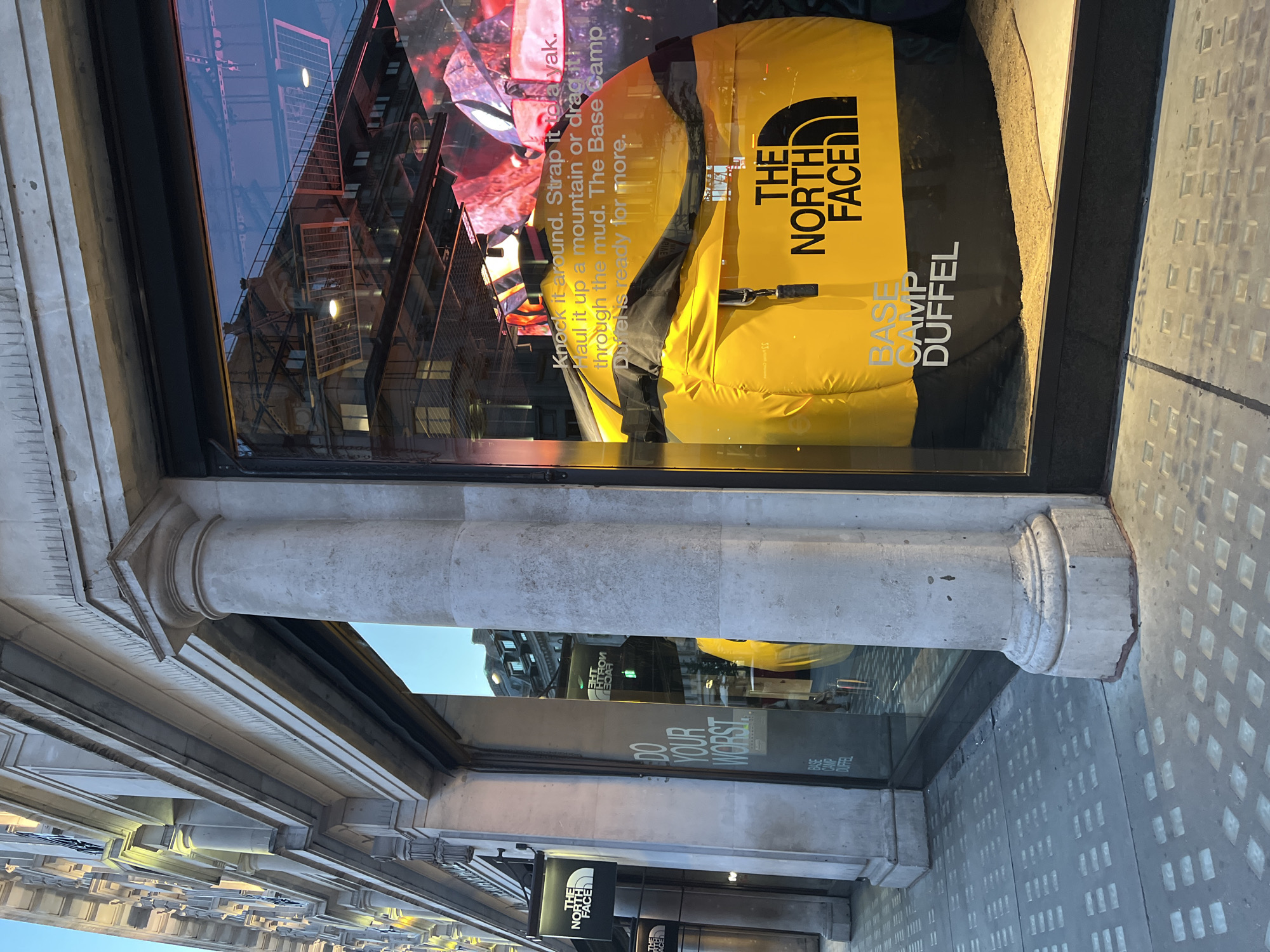 The image shows the exterior of a storefront for The North Face, a retail clothing and outdoor equipment company. The storefront features large glass windows displaying merchandise. Inside the window, a large, bright yellow duffel bag with the "The North Face" logo prominently displayed is the main focal point. The bag is labeled "Base Camp Duffel". Text on the window reads "DO YOUR WORST" and a promotional slogan for the duffel bag is visible: "Knock it around. Strap it to a yak. Haul it up a mountain or drag it through the mud. The Base Camp Duffels ready for more." The building's facade is made of stone with classical architectural elements like columns. The sidewalk in front of the store is paved with patterned paving stones. Reflections of buildings and lights are visible in the store windows. There are no people visible in the image. The lighting suggests it is either early morning or late evening.