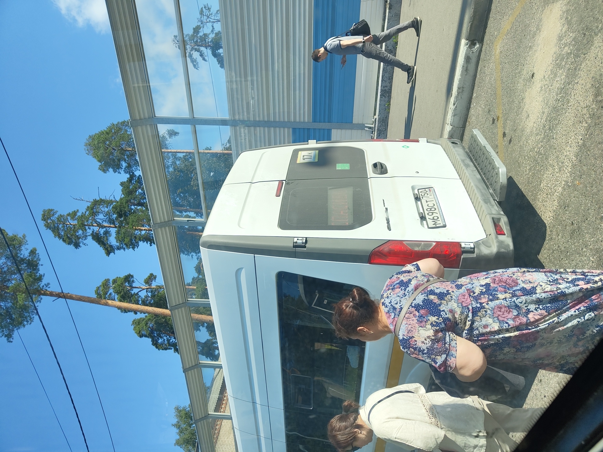 The image depicts a street scene in Zhukovsky, Russia, with a white minibus as the central focus. Two women are standing near the rear of the minibus, one in a cream-colored outfit and the other in a floral dress. A man in a grey t-shirt and pants is walking away from the minibus, looking at his phone. The minibus has a license plate reading "M698CT 750 RUS" and a visible route number "17K". In the background, a sound barrier with reflective panels lines the street, behind which tall pine trees are visible against a bright blue sky. The ground is paved with asphalt and concrete, with yellow markings visible on the asphalt. The lighting suggests it is daytime, likely a sunny day.