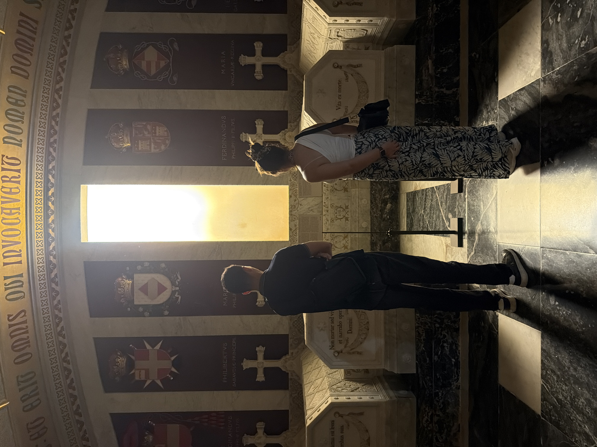 Two individuals, a man and a woman, stand facing away from the camera in a grand mausoleum, likely the Royal Pantheon of San Lorenzo de El Escorial, Spain. The man, on the left, wears a dark blue polo shirt and dark trousers, his arms crossed. The woman, on the right, is dressed in a white one-shoulder top and a black and white patterned skirt, carrying a black bag over her shoulder. They are positioned in front of several ornate sarcophagi. Above the sarcophagi are large panels bearing coats of arms and names, illuminated by a bright light source emanating from a large window or opening at the back of the room. The floor is a striking black and white checkered marble pattern, with significant shadow play. The architectural style suggests a historical and possibly royal burial site, consistent with its location within the Monastery of San Lorenzo de El Escorial.