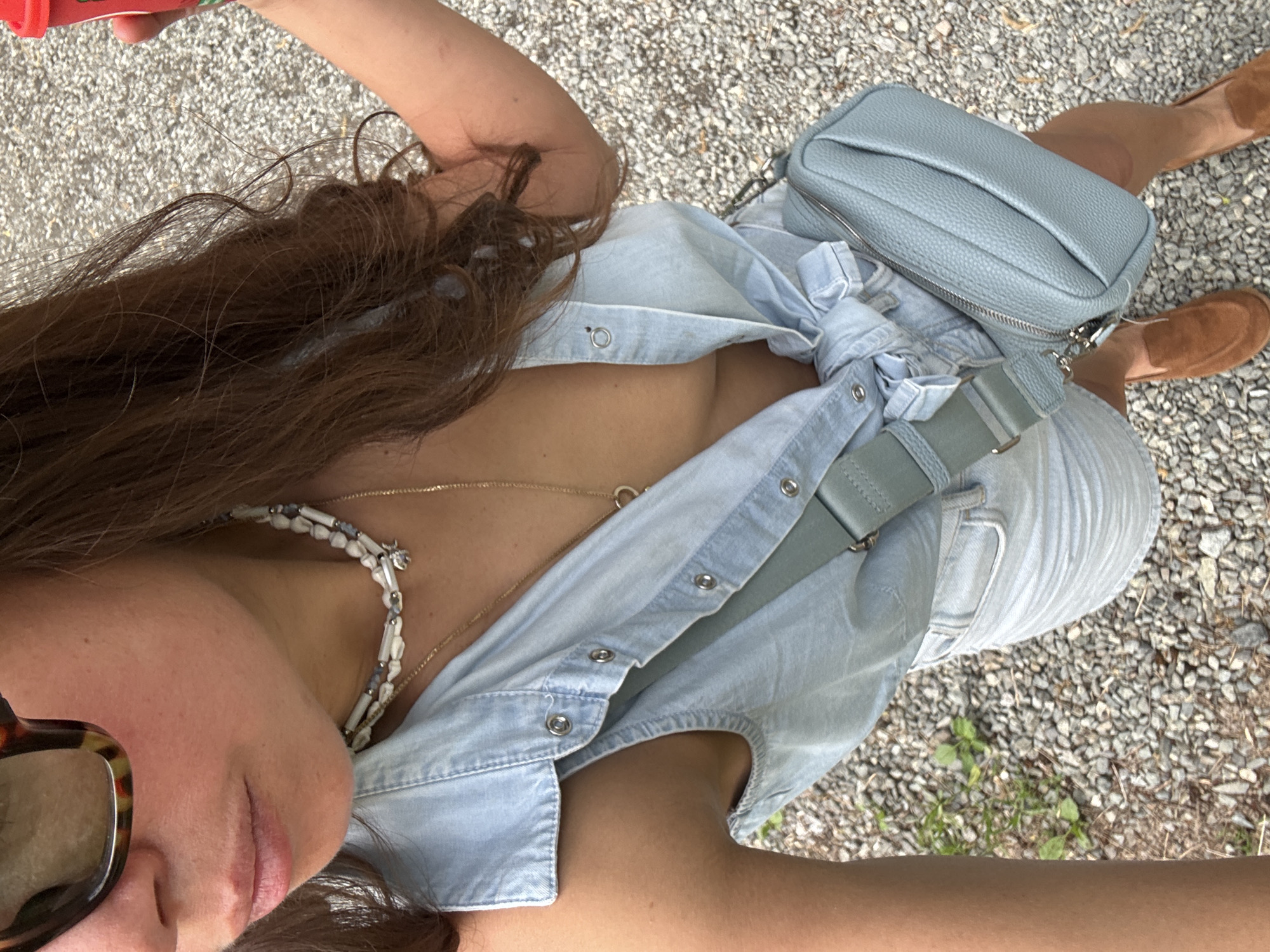 A woman is visible from an overhead perspective, positioned on a gravel surface with sparse vegetation. She has brown hair and is wearing sunglasses. She is also wearing a light blue denim shirt, which is unbuttoned at the top, revealing part of her chest and necklaces. A small light blue purse is positioned across her body, supported by a gray strap. Her feet are visible, wearing brown shoes. A red object is partially visible in the upper left corner. The location is determined to be Sdt Shcherbakovka, Russia.