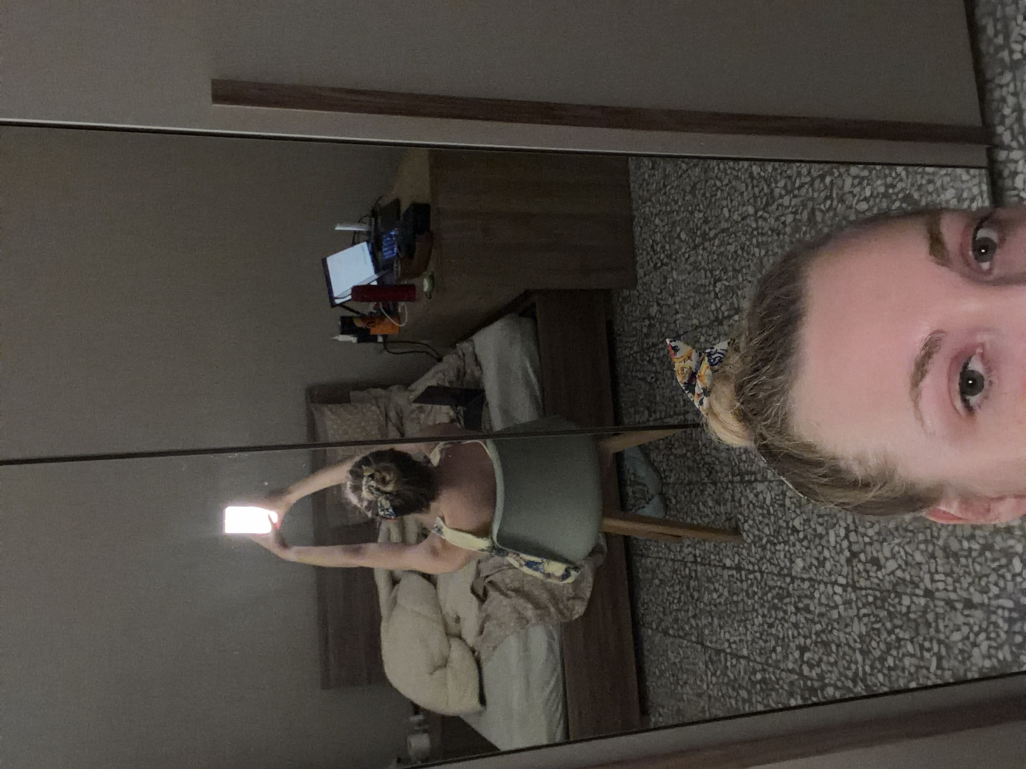 The image shows a reflection in a large mirror of a woman in a bedroom. The woman is sitting in a chair in front of a desk with a laptop. She is holding a phone up to take a selfie. The room appears to be a bedroom with a bed, a nightstand, and a closet. The flooring is tiled. The setting is likely a residential apartment. The image was taken in Catania, Italy.
