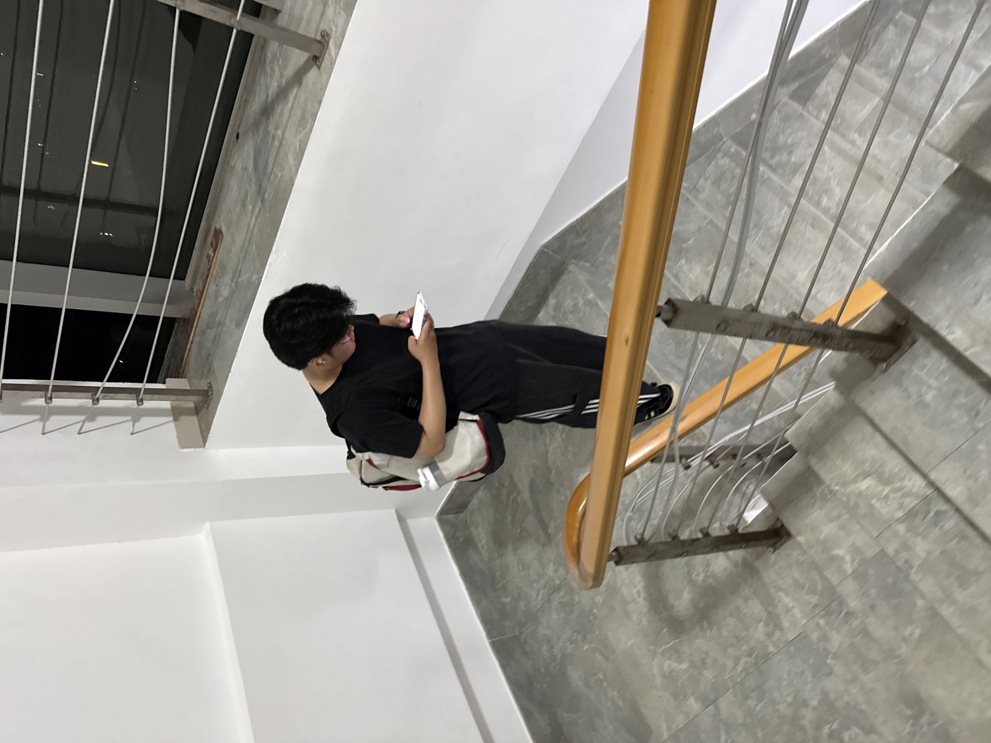 A young person with dark hair, wearing a black t-shirt and black pants with white stripes, stands on a staircase. They have a backpack with white, red, and grey accents slung over their left shoulder and are looking down at a white smartphone in their hands. The staircase has grey tiled flooring and a wooden handrail with metal railings. The background shows a white wall and a reflective surface that appears to be a window or glass railing overlooking a dark area. The scene is indoors, likely in a building in Huizhou, China.