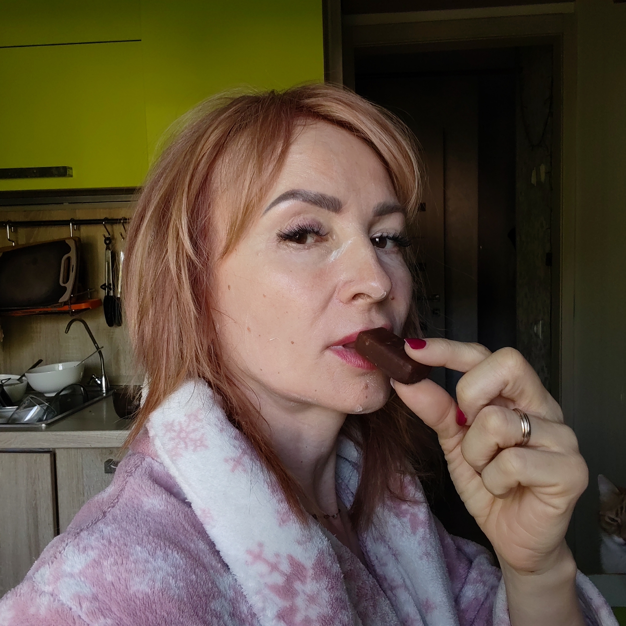 A middle-aged woman with fair skin, reddish-blonde hair, and red nail polish is holding a dark chocolate candy to her mouth. She is wearing a pink and white snowflake-patterned robe. Her hair is slightly messy, and there appears to be a light dusting of powder or flour on her face. She has visible eyelashes.
Behind her is a kitchen setting, with light wood-grained cabinets and a bright yellow wall cabinet. A sink, cookware, and utensils are visible on the counter. Behind the woman, a dark doorway is visible, with a glimpse of an orange tabby cat to the right.
The image was taken in the city of Chaikovsky, Russia.