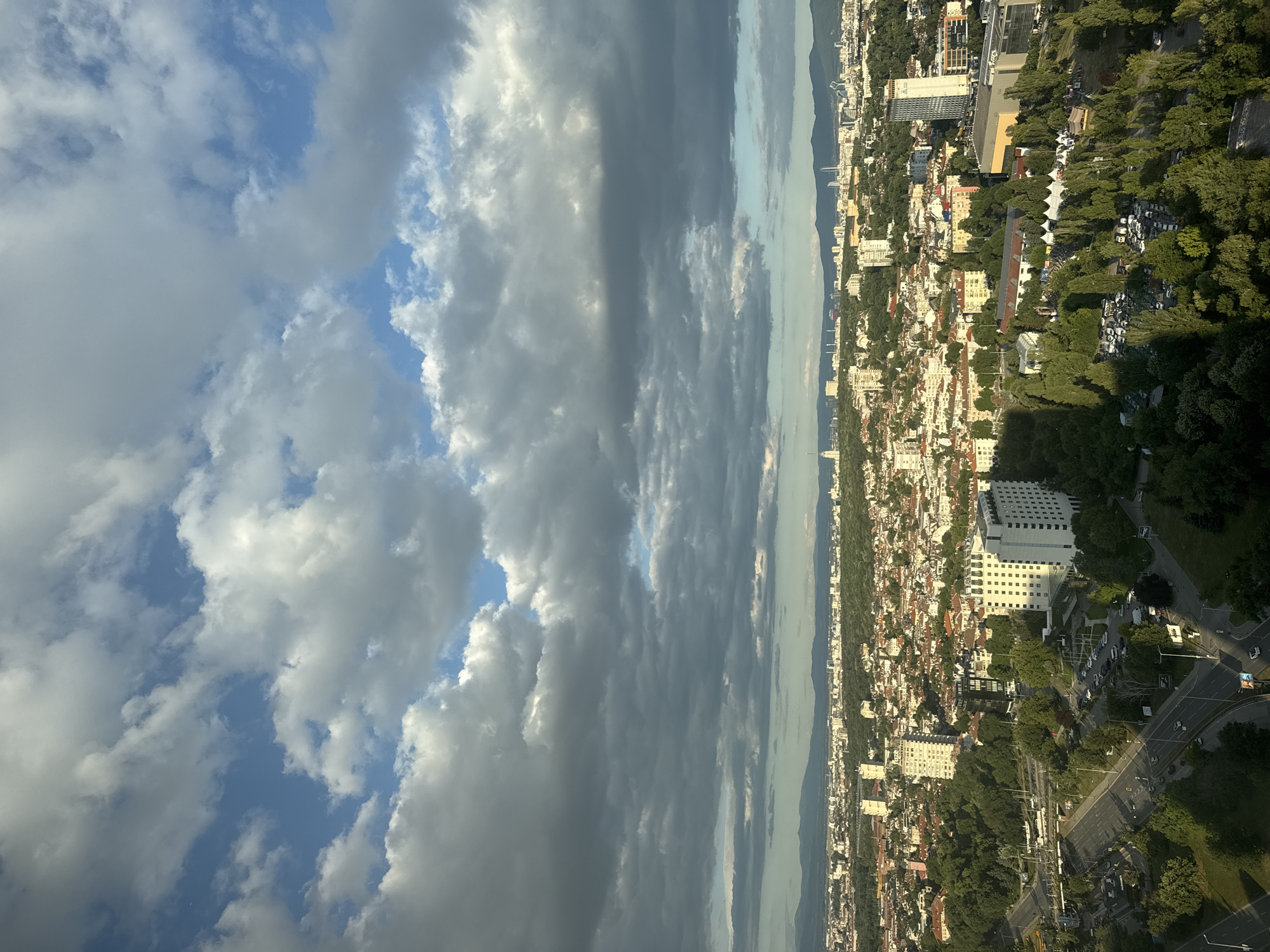 The image presents an aerial view of a cityscape, likely Sofia, Bulgaria, under a dramatic sky filled with a mix of light and dark clouds. In the foreground, a busy road with cars navigates through a green, tree-filled area. A prominent white hotel-like building with numerous windows stands out, adjacent to a parking lot. Further into the city, a dense urban landscape unfolds, characterized by a mosaic of buildings with red-tiled roofs and taller, modern structures. The distant horizon is lined with mountains, softened by the atmospheric conditions. The overall impression is one of a sprawling city bathed in the diffused light of an overcast, yet potentially clearing, day.