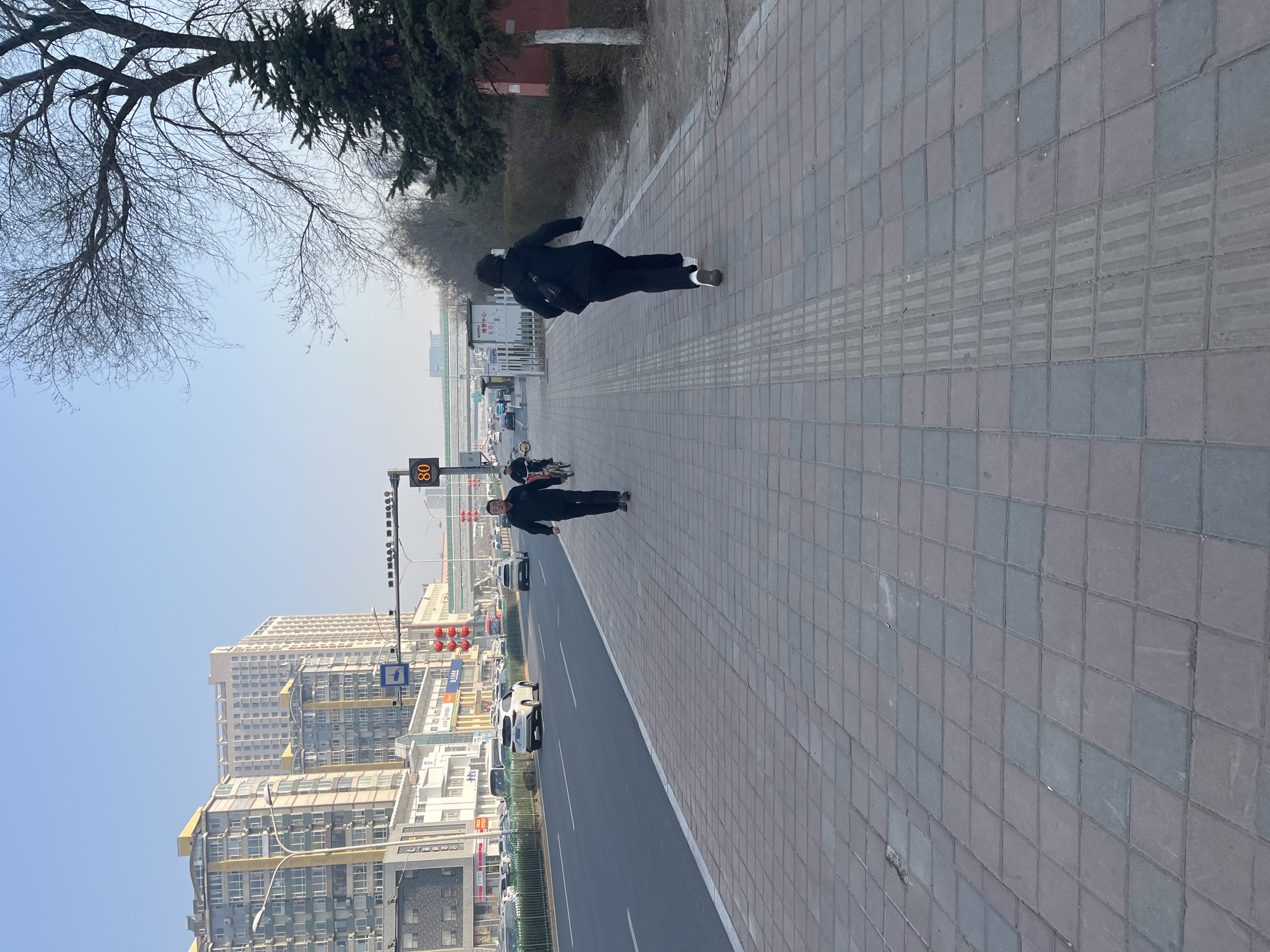 Two people are walking on a wide sidewalk in Changchun City, China. The sidewalk is made of gray paving stones with a strip of tactile paving for the visually impaired. One person is walking ahead of the other. They are both dressed in dark clothing. The street next to the sidewalk has multiple lanes of traffic with cars. Tall buildings are visible in the background. The sky is light blue. A traffic light with a digital speed limit display is visible. Bare tree branches and evergreen trees are in the upper left corner.