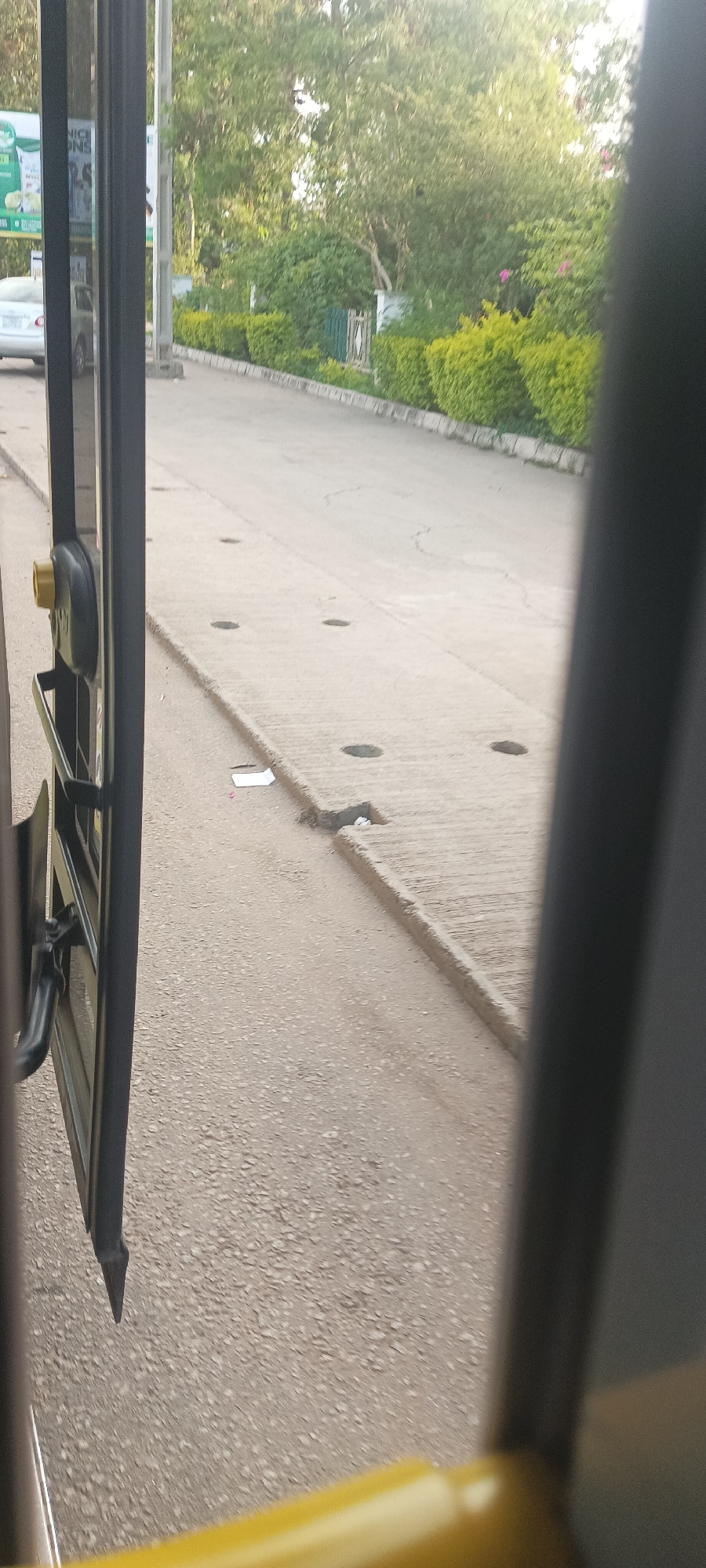 The image presents an outdoor scene viewed from within a vehicle, likely a bus, through its partially open door. A yellow handrail is visible in the lower right foreground, indicating the interior of the vehicle.
Outside, a paved road or pathway stretches into the distance. This pathway features a distinct section of concrete slabs with several circular holes, possibly for drainage or utility access. The road surface shows some cracks, and a piece of white litter is visible on the ground near the foreground.
To the left, part of a white or silver sedan is visible, suggesting another vehicle or a parked car. Beyond the immediate paved area, lush green hedges line what appears to be a curb or low wall, separating the road from dense trees and foliage. A small gate or fence is discernible amidst the greenery further back.
In the background, natural light suggests it is daytime, with clear weather. On a billboard or sign in the far background, partially obscured by the vehicle's frame and trees, fragments of text such as "NICE," "ONS," and possibly "OOL" can be faintly made out, but are too blurry to be fully legible. No people are visible, and no specific activity beyond observing the passing or stationary scene is depicted. The overall setting is green and appears to be in a natural or semi-urban environment.