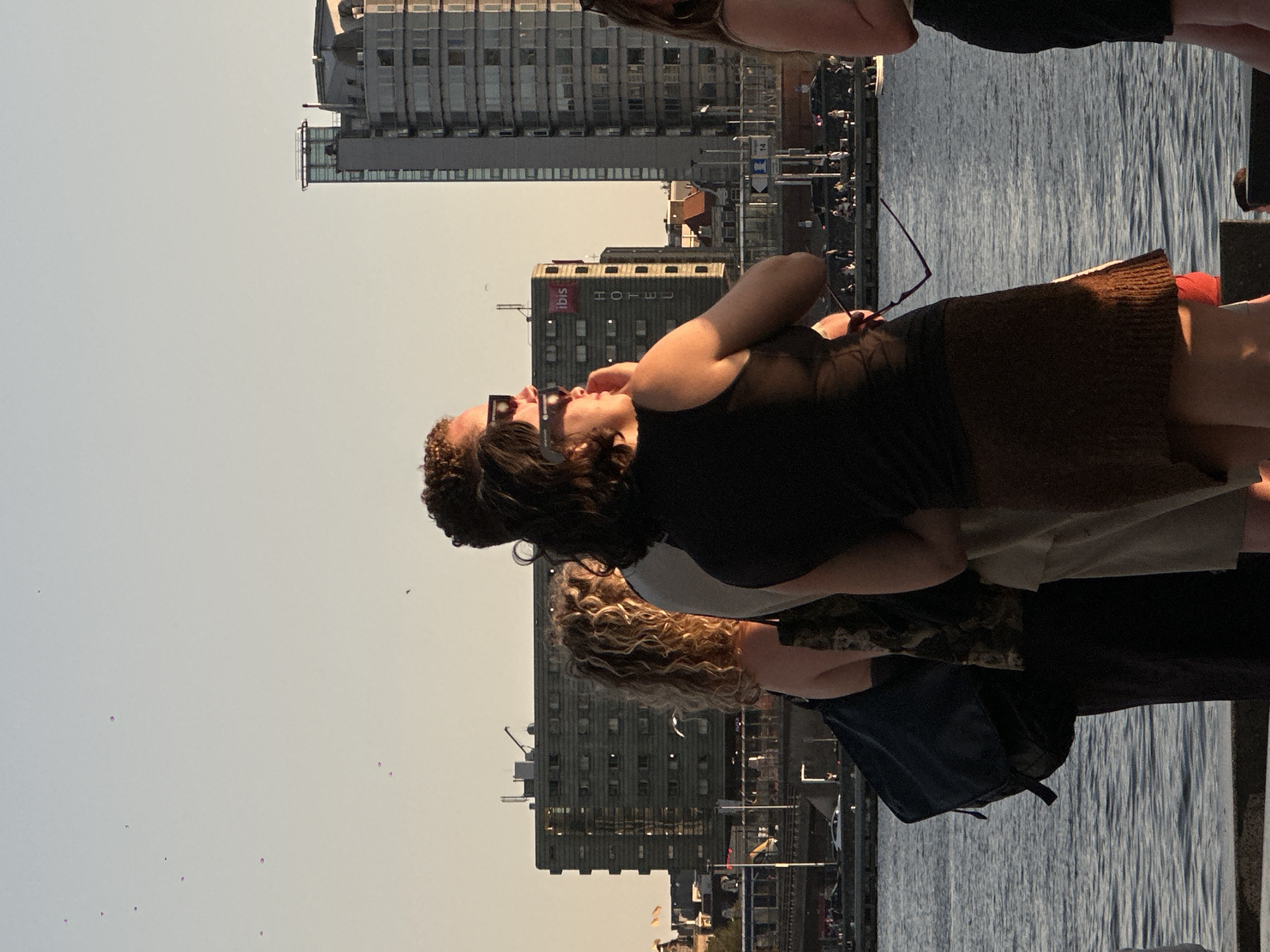 The image shows a group of four people standing by a waterfront in Amsterdam. The weather appears to be clear and sunny, with the golden hour light casting long shadows. In the background, there are several buildings, including a tall modern building and a mid-rise hotel with the "ibis HOTEL" sign visible. The waterfront is lined with a railing, and beyond that, there is a street with cars and pedestrians. The water is calm, reflecting the light and the surrounding buildings. The people in the foreground are casually dressed, and some are wearing sunglasses. One person is holding a pair of sunglasses. The overall scene is a snapshot of urban life in Amsterdam during a pleasant evening.