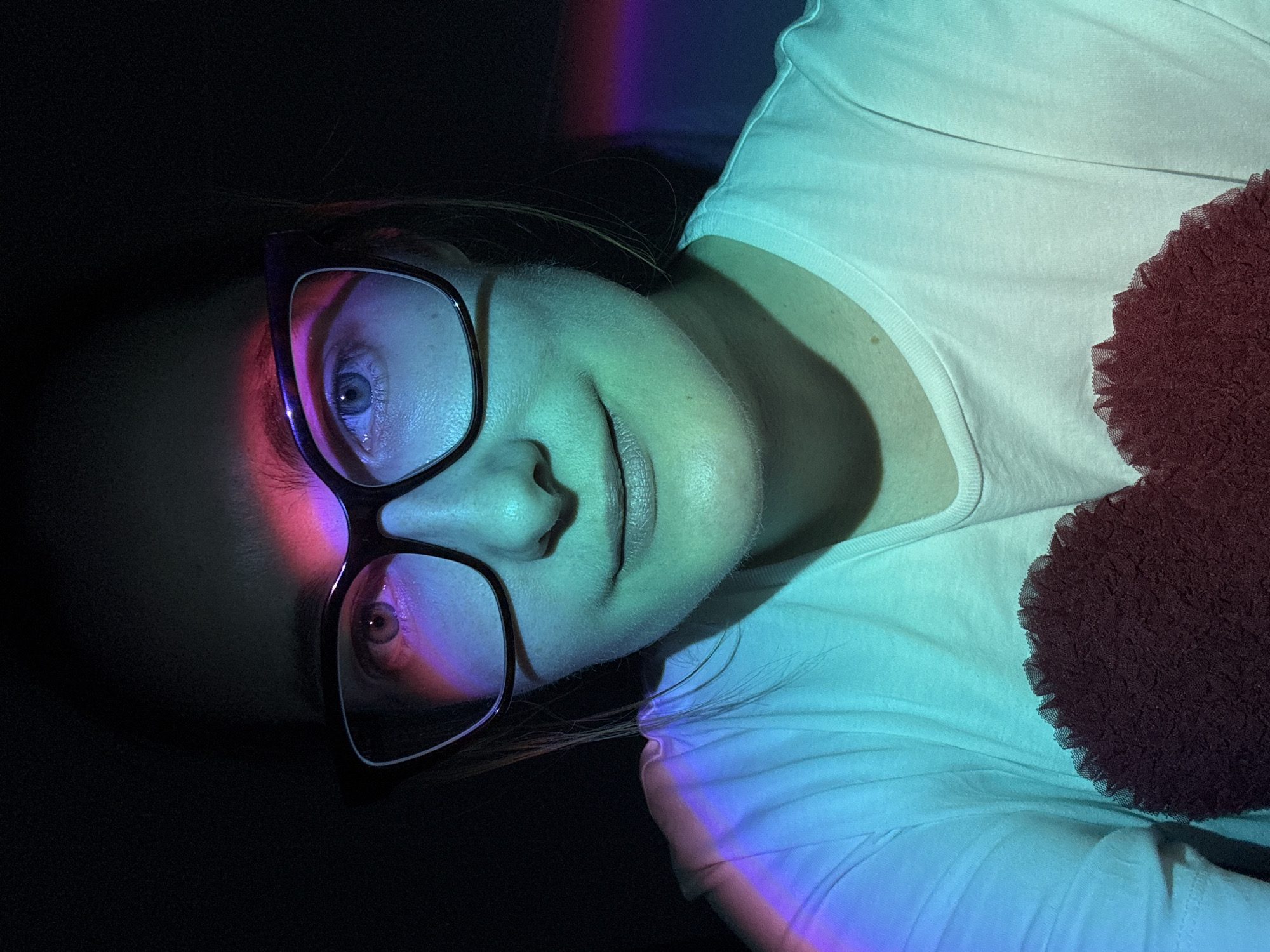 A portrait of a woman with glasses is illuminated with cyan, pink and purple light. The woman is wearing a white t-shirt and appears to be reclining, with a fuzzy, dark brown object positioned near her torso. The woman is wearing large, dark-rimmed glasses. The scene is generally dark, with illumination provided only by colored lights. The scene is unlikely to be Riga, Latvia due to the absence of any location-identifiable indicators.