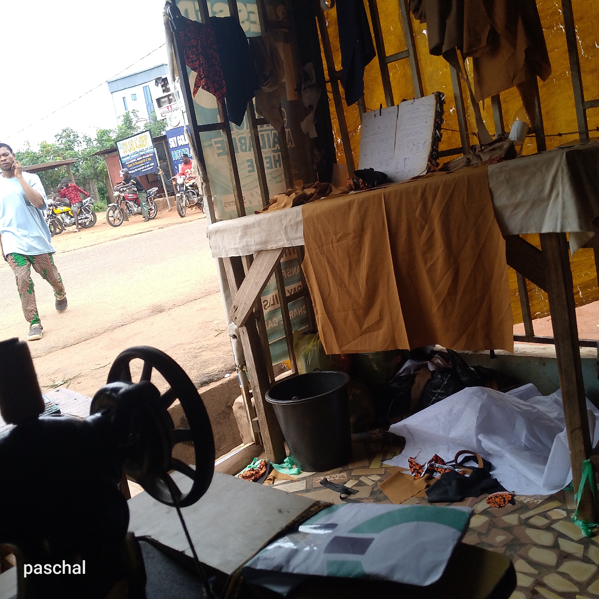 The image depicts a street scene in Nsugpe, Nigeria, captured from inside a tailor's shop. A man is walking on the street while using a mobile phone. Three men on motorcycles are parked nearby. The tailor's shop is cluttered, with fabrics, a bucket, and a sewing machine. A table covered with fabric and other materials is also visible. There are signs visible in the background, one reading "SKY GOLD HOTEL" and another partially visible sign reading "SKYLAND ACADEMY Close To Descent Oil & Gas".