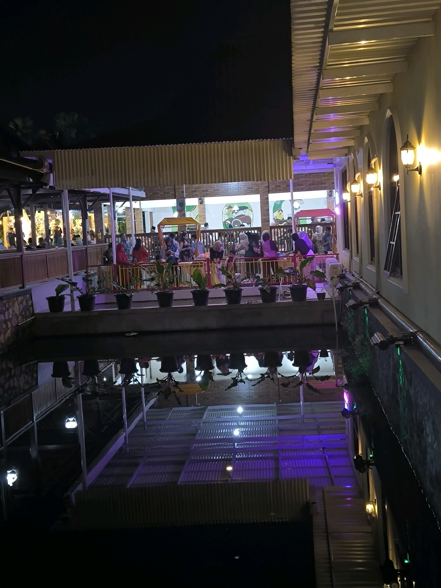 The image depicts a nighttime scene at an outdoor seating area, possibly a restaurant or cafe, with a reflective water feature in the foreground. The area is illuminated by warm-toned wall sconces and purple lights, creating a vibrant atmosphere. Several potted plants are arranged along the edge of the elevated seating area, which is populated by groups of people enjoying their evening. The architecture features corrugated metal roofing and a wall with arched windows. The water feature reflects the lights and structures above, adding depth and visual interest to the scene.