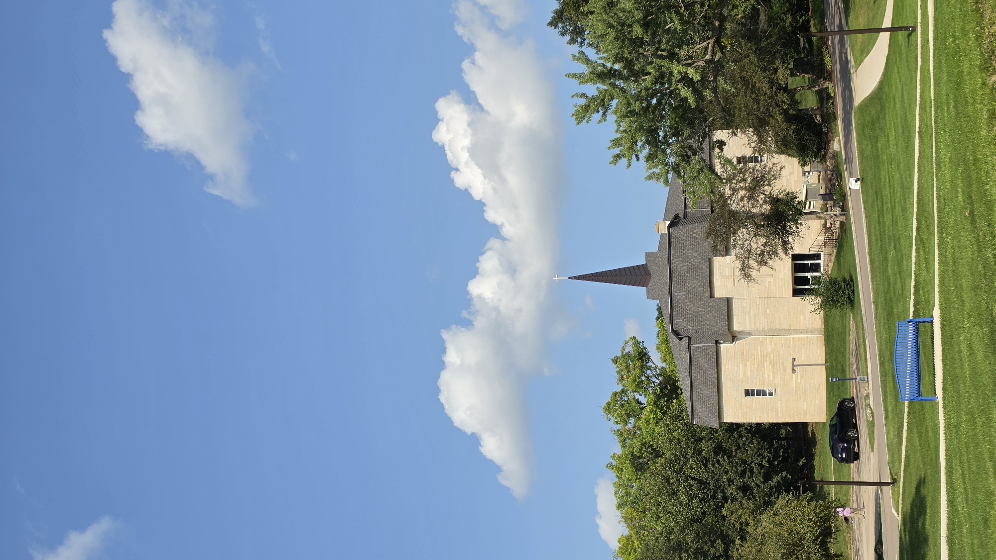 An outdoor, eye-level, vertical shot captures a building that appears to be a church or similar institution. The structure is made of light-colored stone and features a dark, pointed steeple topped with a white cross. Lush green trees and grass surround the building, suggesting a natural or park-like setting. The sky is a vibrant blue with scattered white clouds. In the foreground, a bright blue park bench sits on a well-maintained lawn. A paved walkway curves through the grassy area, leading towards the building. A dark-colored car is parked on a paved area near the building. A single person, wearing a pink shirt and dark pants, walks away from the viewer near the car. The scene is illuminated by bright sunlight, casting subtle shadows. The location is identified as Elgin, United States of America.