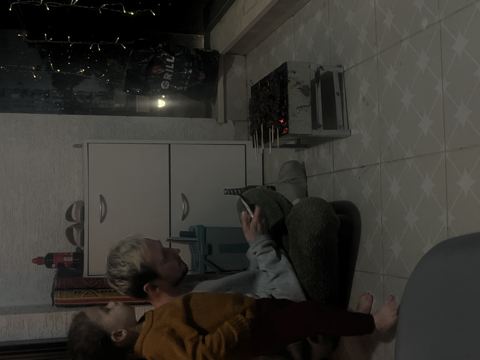 The scene is set on a tiled balcony or patio at night in Kiryat Bialik, Israel. An adult male and a young child are present.
The adult male, with blond hair, is seated cross-legged on the patterned light-colored tiled floor. He is wearing a light grey long-sleeved top and dark grey trousers, and is barefoot. He holds a white smartphone in both hands, looking down at the screen. To his left stands a young child, barefoot, wearing a mustard yellow long-sleeved shirt and dark trousers. The child is facing right, observing the area near the adult.
In front of the seated male is a small, rectangular, metallic charcoal grill with hot embers and several skewers of meat cooking. A dark glass bottle is on the floor near the grill. Behind the male, against a light-colored textured wall, is a two-drawer white cabinet with silver handles. On top of the cabinet are a red-capped bottle and a pair of light-colored slip-on shoes. A patterned textile is partially visible behind these items. A light green plastic container sits between the male and the cabinet.
To the right of the scene, a large glass door or window looks out into darkness. A black bag labeled "GRILL" is placed on the ground near this window, and vertical strings of yellow-orange fairy lights are visible outside. Artificial lighting illuminates the immediate area.
