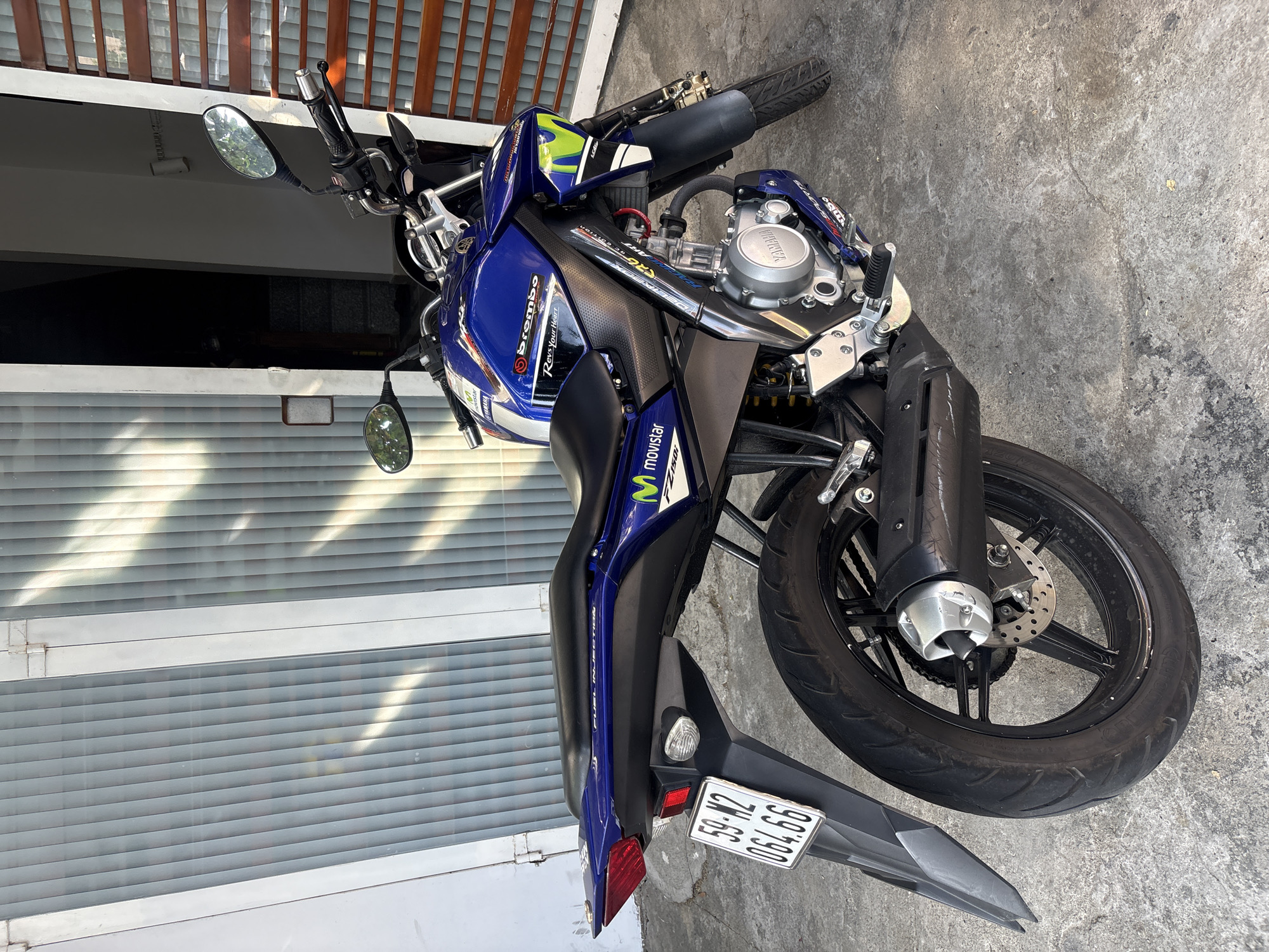 The image shows a blue and black Yamaha motorcycle parked on a concrete surface next to a building with a roll-up door and slatted wooden accents above. The motorcycle has visible branding including "Brembo" and "Movistar". The license plate is partially visible, displaying "59-N2 006.66". The location is Nha Trang Ward, Vietnam.