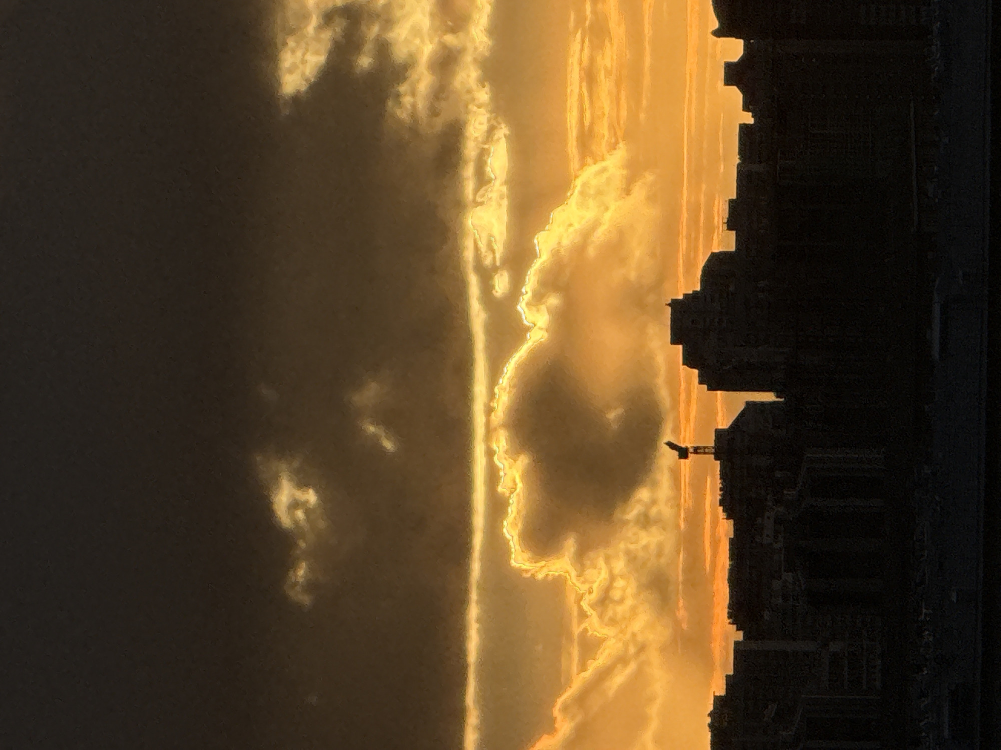 The image depicts a sunset over a cityscape, likely in Moscow, Russia. Silhouetted buildings are visible on the right side of the frame. A construction crane is visible amidst the buildings. The sky is filled with golden clouds, and the sun appears to be setting, casting a warm glow across the scene. The left side of the image is darker, possibly due to a storm cloud.