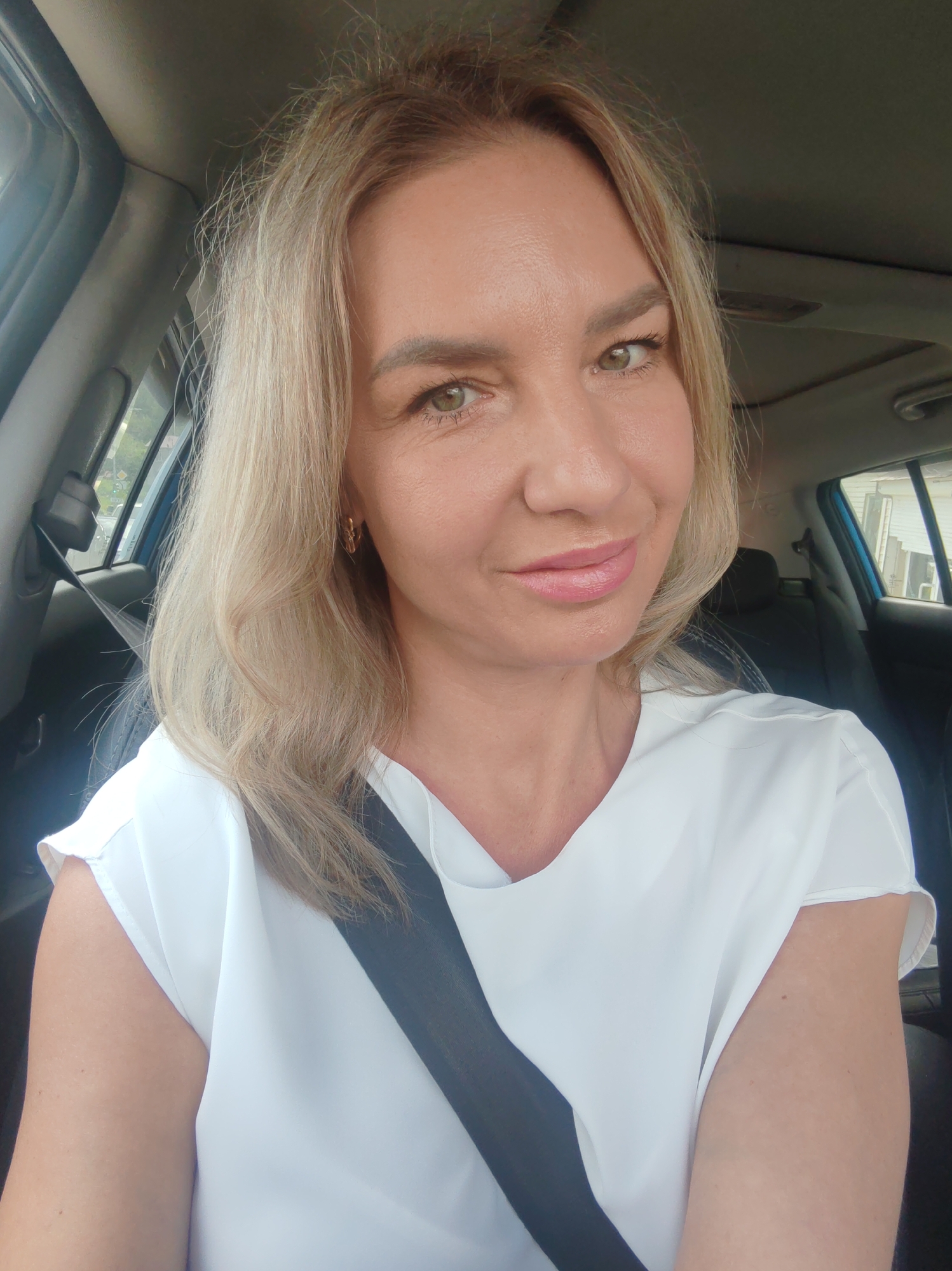 A medium shot selfie shows a Caucasian woman with blonde hair and green eyes, wearing a white v-neck top and a black seatbelt, looking directly at the camera with a slight smile. She appears to be in the passenger seat of a car, indicated by the car interior visible in the background. The woman has light makeup, including pink lipstick and eye shadow. The overall lighting suggests it is daytime. The image does not contain enough information to determine the specific location within Petropavlovsk-Kamchatsky, Russia.