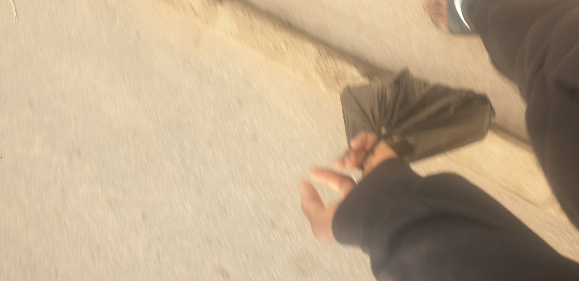 A person's hand, wearing a black hoodie, is holding a black plastic bag as they walk on a paved surface. The image is taken from a first-person perspective and is blurry due to motion. The environment appears to be outdoors, possibly a sidewalk or road, with a light-colored ground. No specific landmarks of Jos, Nigeria are visible.