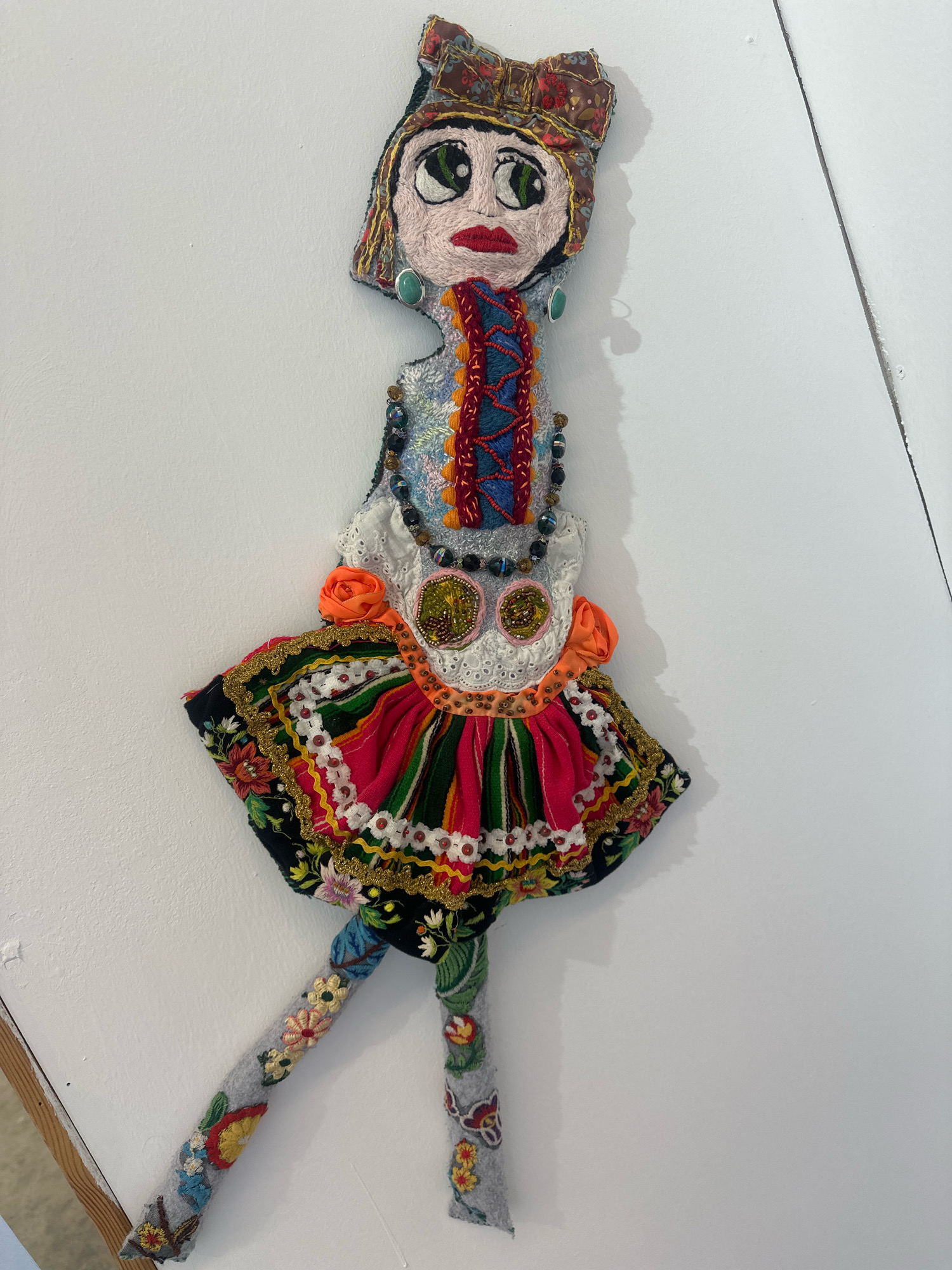 The image displays a handmade textile doll hanging on a white wall, likely indoors. The doll is intricately decorated with various fabrics, embroidery, beads, and sequins, suggesting a folk art or artistic creation. It has a detailed face with large green eyes, red lips, and a patterned headscarf or hat. The doll's body is adorned with a colorful, layered dress that appears to be made of striped and floral fabrics, embellished with gold trim and small orange rosettes. Its limbs are also made of fabric, with embroidered floral patterns. The doll is positioned in the center of the frame, casting a shadow to its right. The white wall serves as a neutral background, with a faint diagonal line suggesting a joint or seam in the wall or a light fixture. No specific location in Mate Asher Regional Council, Israel, can be identified from the image itself.