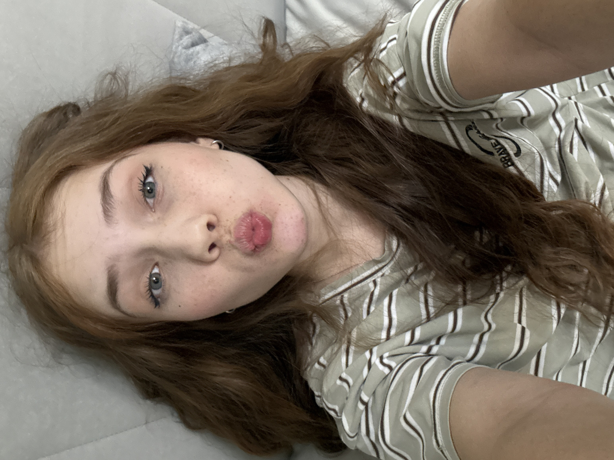A young female is depicted in a selfie, lying on a light-colored surface, likely a bed or couch, in an indoor setting. She has long, wavy reddish-brown hair, light blue or grey eyes with visible mascara, and fair skin with freckles across her nose and cheeks. Her lips are formed into a playful pout.
She is wearing a light green or beige t-shirt with thin vertical stripes in white and brown. The word "BRAVE" is visible in black text on the shoulder area of her shirt. A silver hoop earring is seen in her visible left ear. The image is taken from a slightly elevated perspective, typical of a selfie. No specific time of day or weather conditions are discernible, nor are any direct location cues indicating Naberezhnye Chelny, Russia, visible within the image. The overall impression is one of relaxation and playfulness.